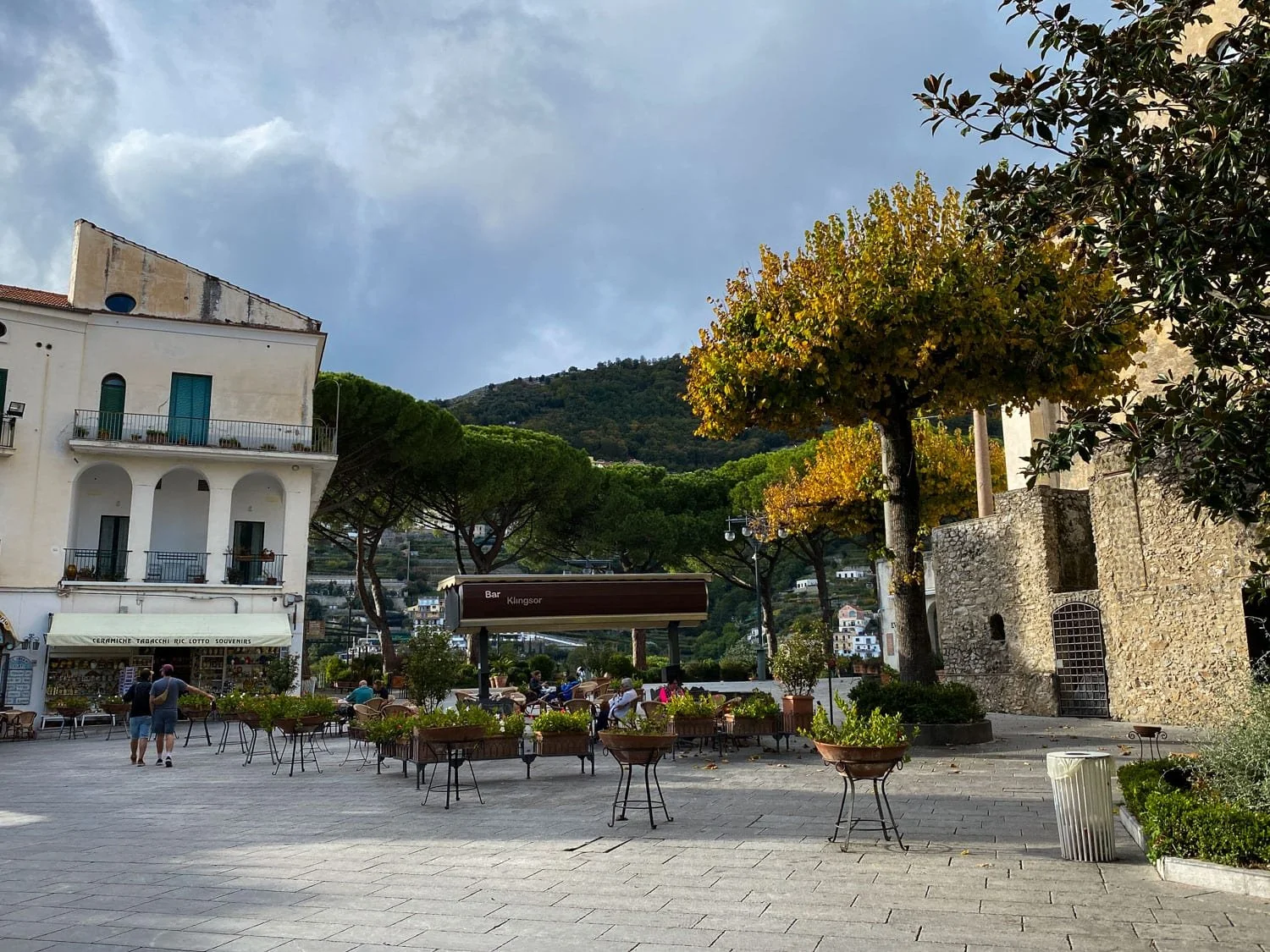 Open piazza in Ravello lined with cafés, potted plants, and historic stone buildings beneath dark storm clouds. Visitors sit outdoors surrounded by mountain views and umbrella pine trees.