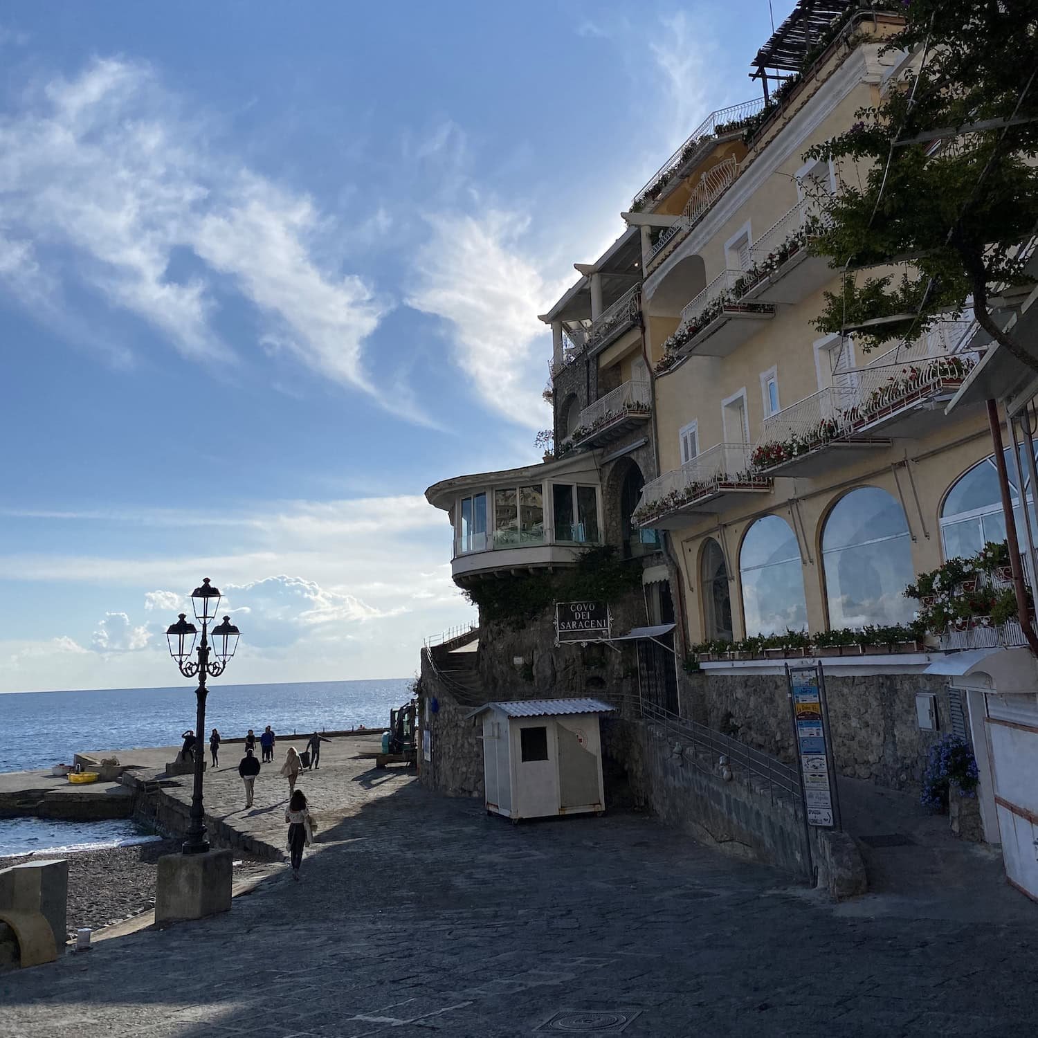Seaside promenade in Amalfi with people walking along a stone path beside the ocean and pastel buildings rising above. The location shows the convenience of staying close to the waterfront in Amalfi.