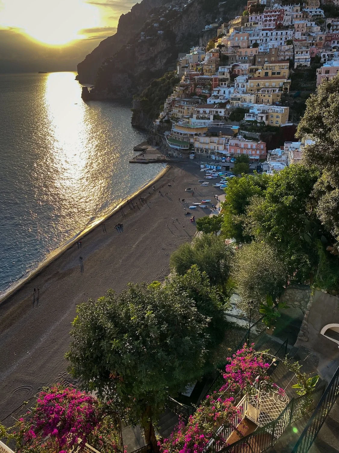 Sunset view over Positano beach with golden light reflecting on the water. Colorful cliffside buildings rise above the shoreline on the Amalfi Coast surrounded by green trees and bright pink flowers.
