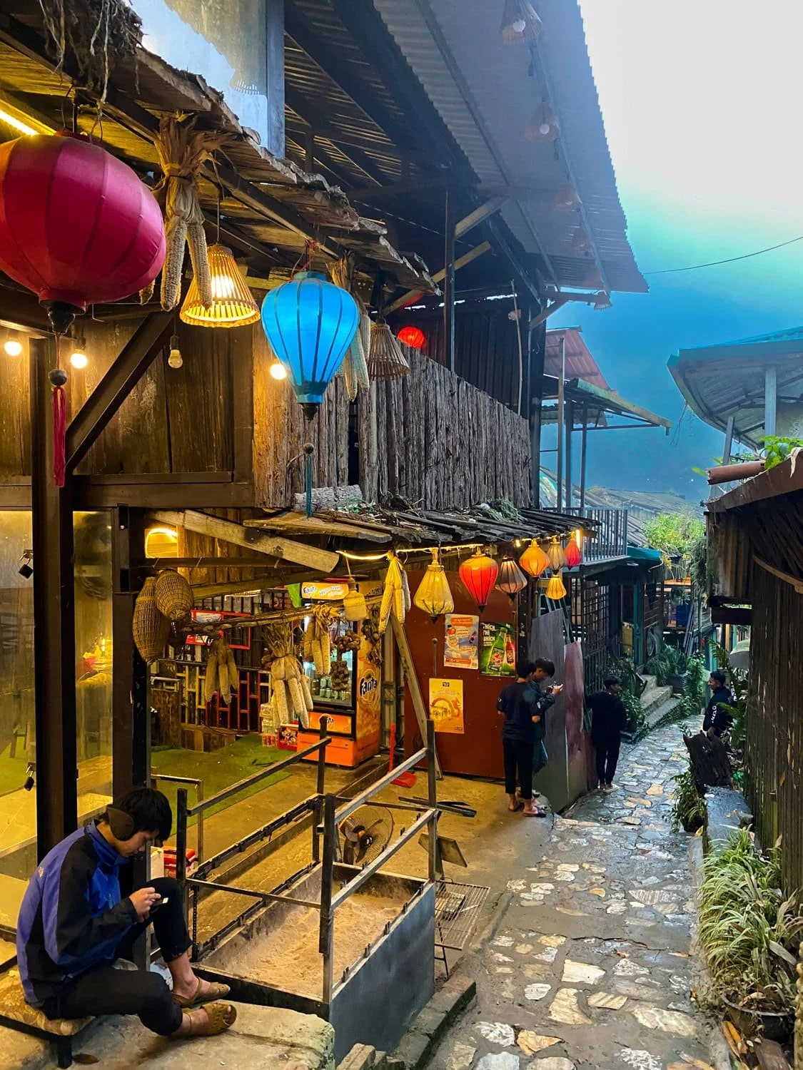 A narrow alleyway in Cat Cat Village lined with shops and hanging lanterns, with a few locals relaxing along the stone path at dusk.