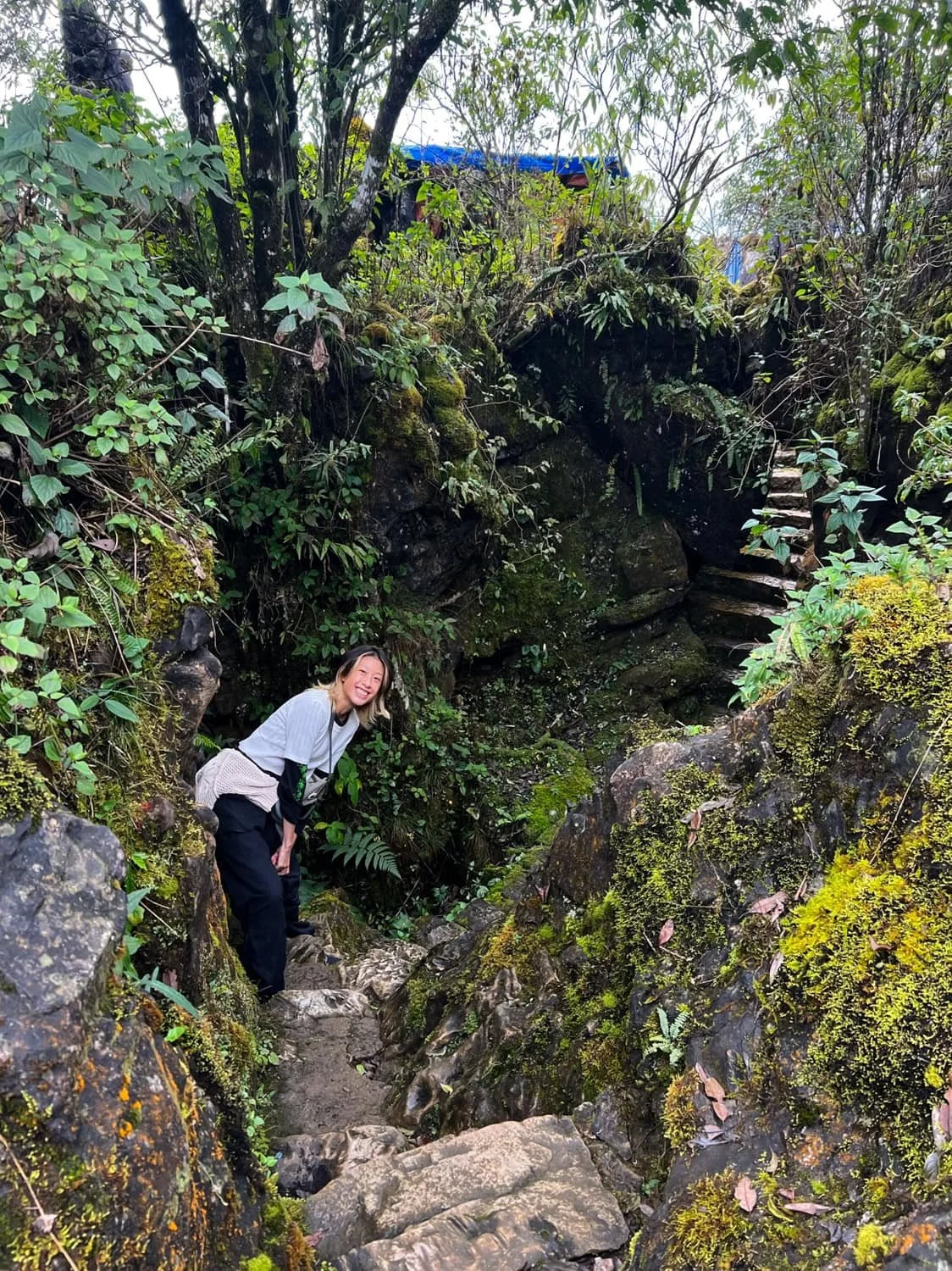 A smiling woman stands in a mossy, rocky forest path surrounded by dense vegetation and large stones in Sapa. Serene hiking spots like this suggest that Sapa is worth visiting for outdoor adventurers.