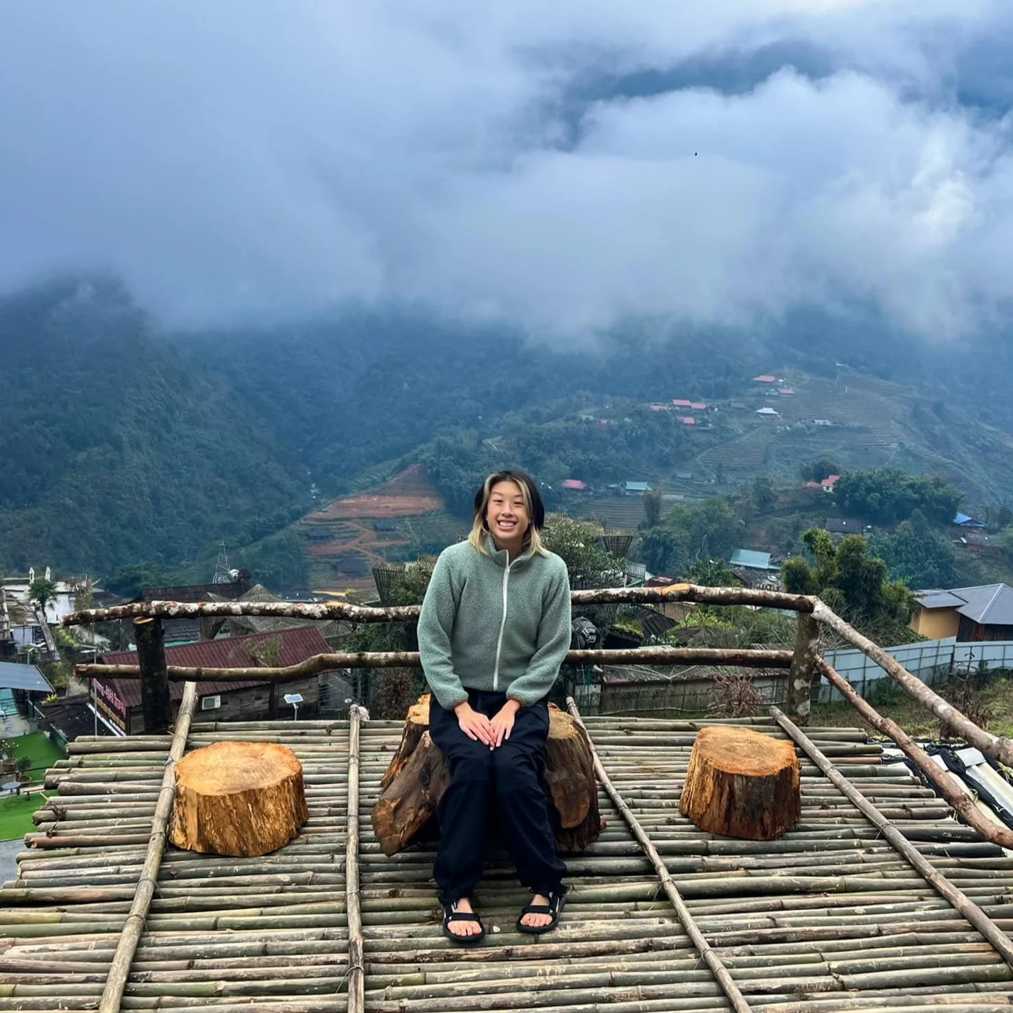A woman sits on a bamboo terrace with log stools, smiling against a scenic backdrop of rice terraces and mountain valleys in Cat Cat Village, partially covered in mist.