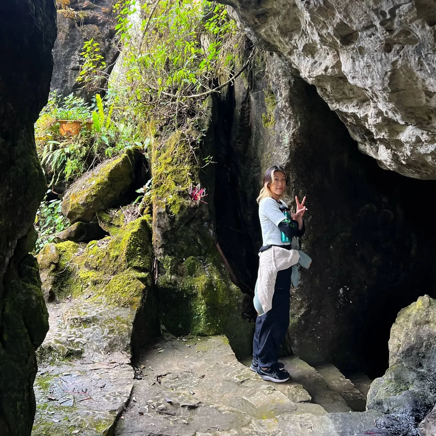 A woman flashing a peace sign while standing at the entrance of a moss-covered cave surrounded by jungle greenery and rugged stone in Sapa. Unique nature spots like this often convince visitors that Sapa is worth visiting.