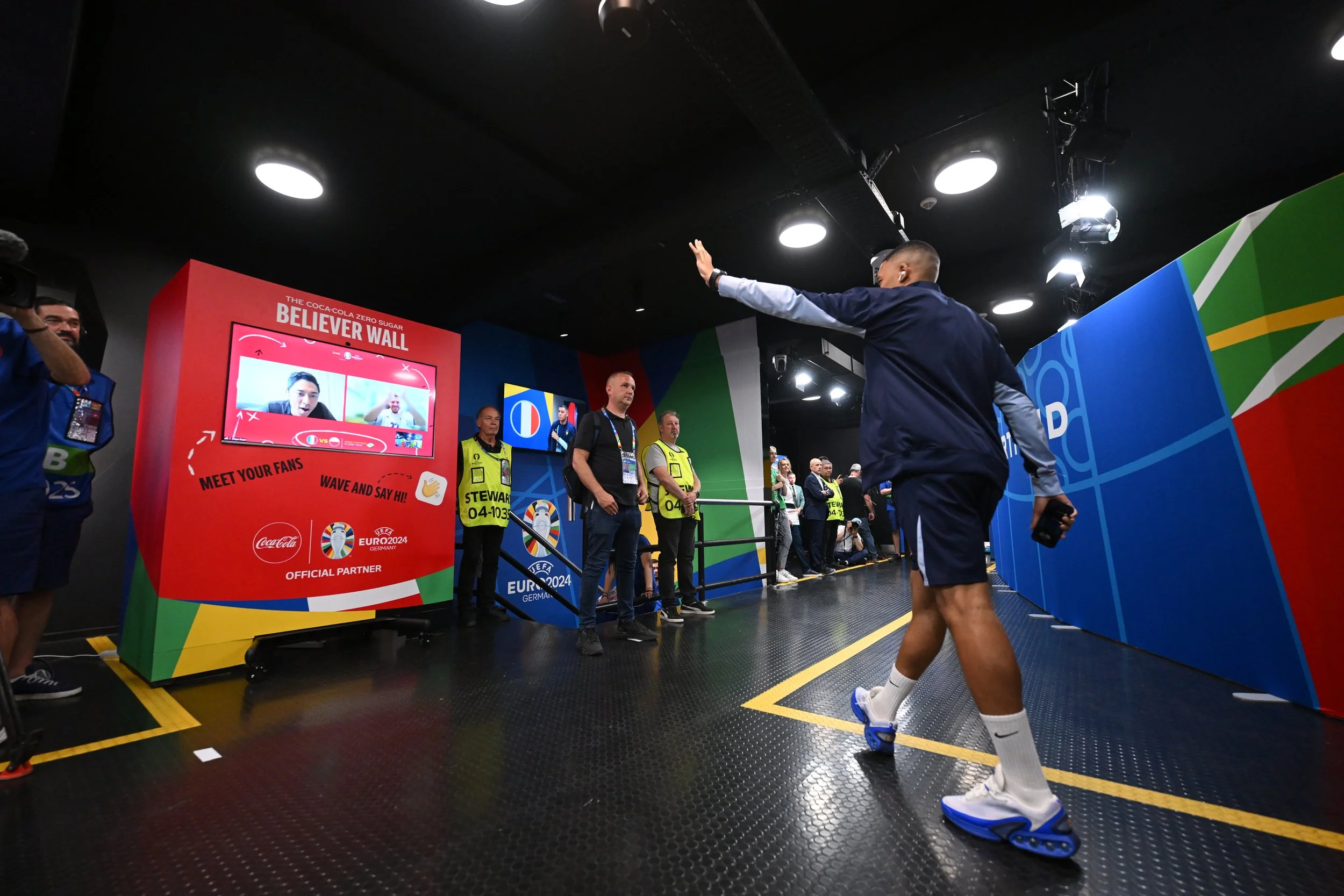 A football player dressed in a navy jacket, shorts, and sandals is greeting fans at an indoor event area decorated with colorful panels and a large Euro 2024 banner. Several officials and spectators watch as he waves and extends his hand.