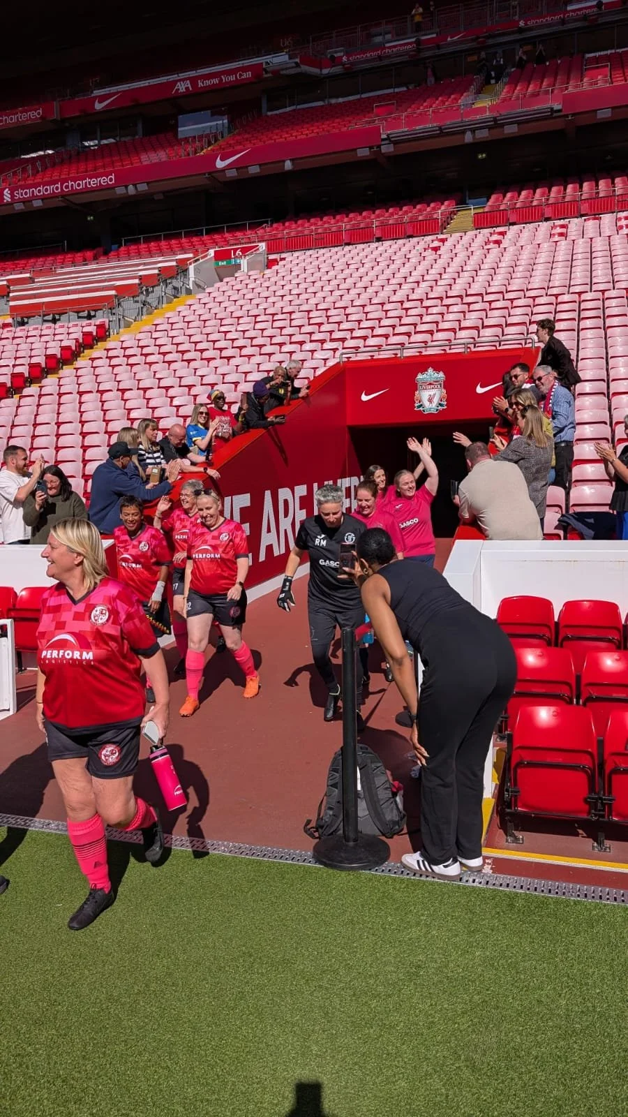 Liverpool football players and staff walking through a stadium tunnel onto the field, with fans clapping and cheering.