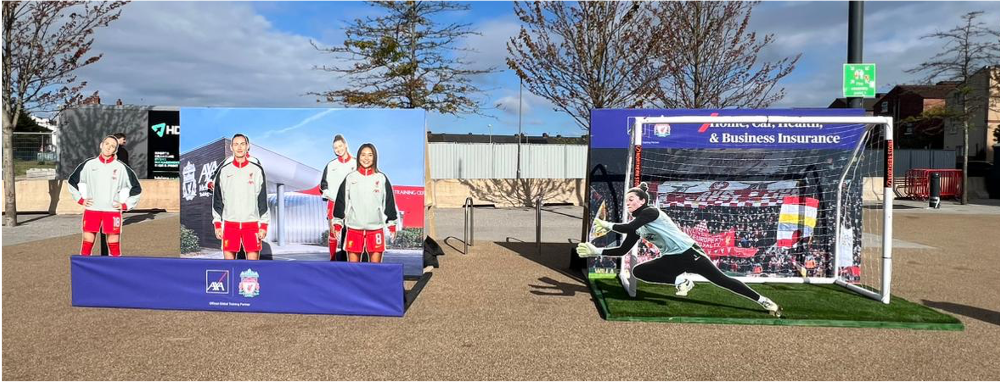 Outdoor soccer-themed photo featuring a female goalkeeper diving to save a shot, with a net and back wall displaying a Liverpool FC banner, and cardboard cutouts of female soccer players in Liverpool uniforms.
