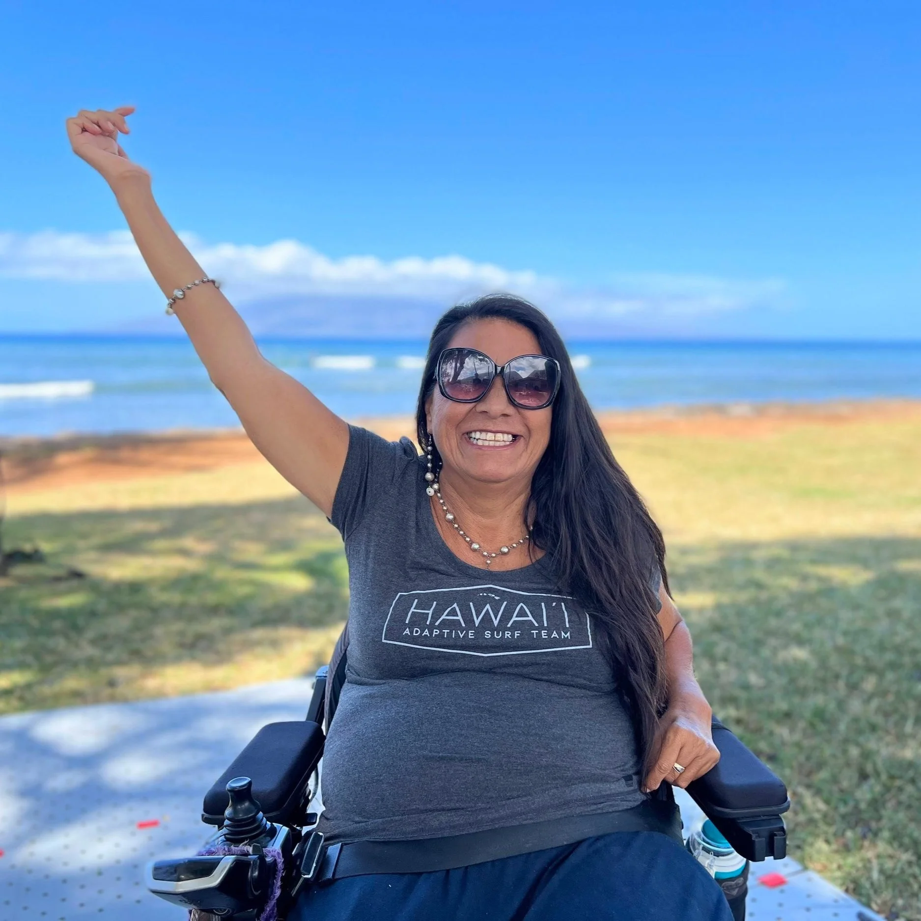 Woman sits in a wheelchair with her right arm raised and a smile on her face. She is wearing a grey scoop neck tee shirt with a white logo on the front that says Hawaii Adaptive Surf Team