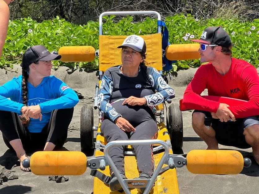 A woman in blue sits to the left of a woman who is quadrapelegic in a beach wheelchair. A man sits to her right. both people are looking to the center at the woman in the beach wheelchair.