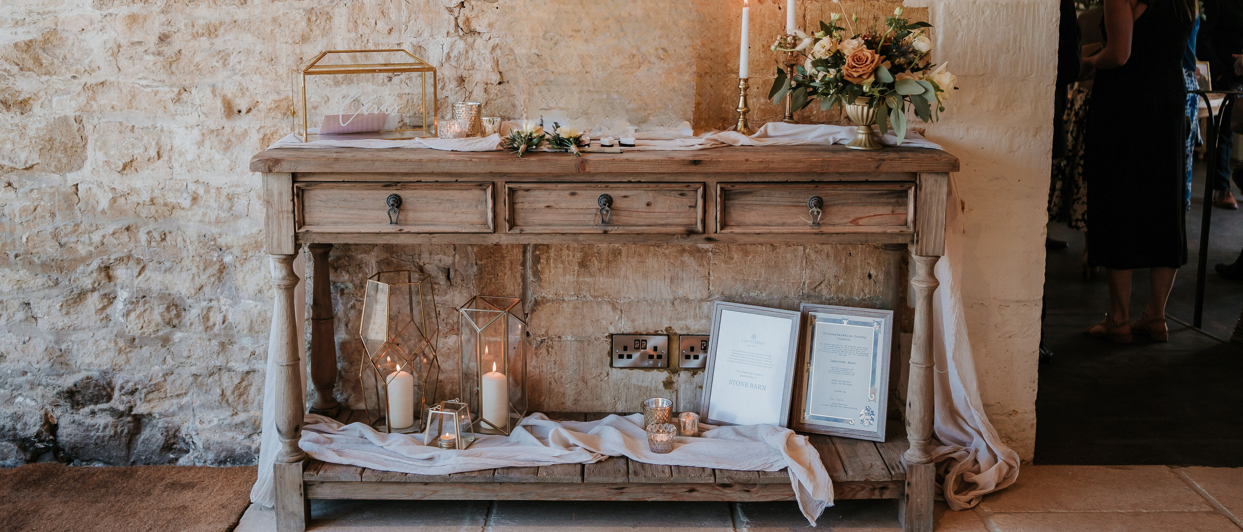 A wedding guestbook and cards display table set up at Lapstone Barn. The table is styled in a rustic theme with layered gauze runners, neutral florals and gold and brass decorative accents. The setup has a soft, elegant countryside feel.