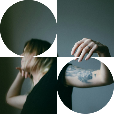 A person with blonde hair wearing a black shirt, showing a tattoo on their arm, adjusting their hair with a blurred background.