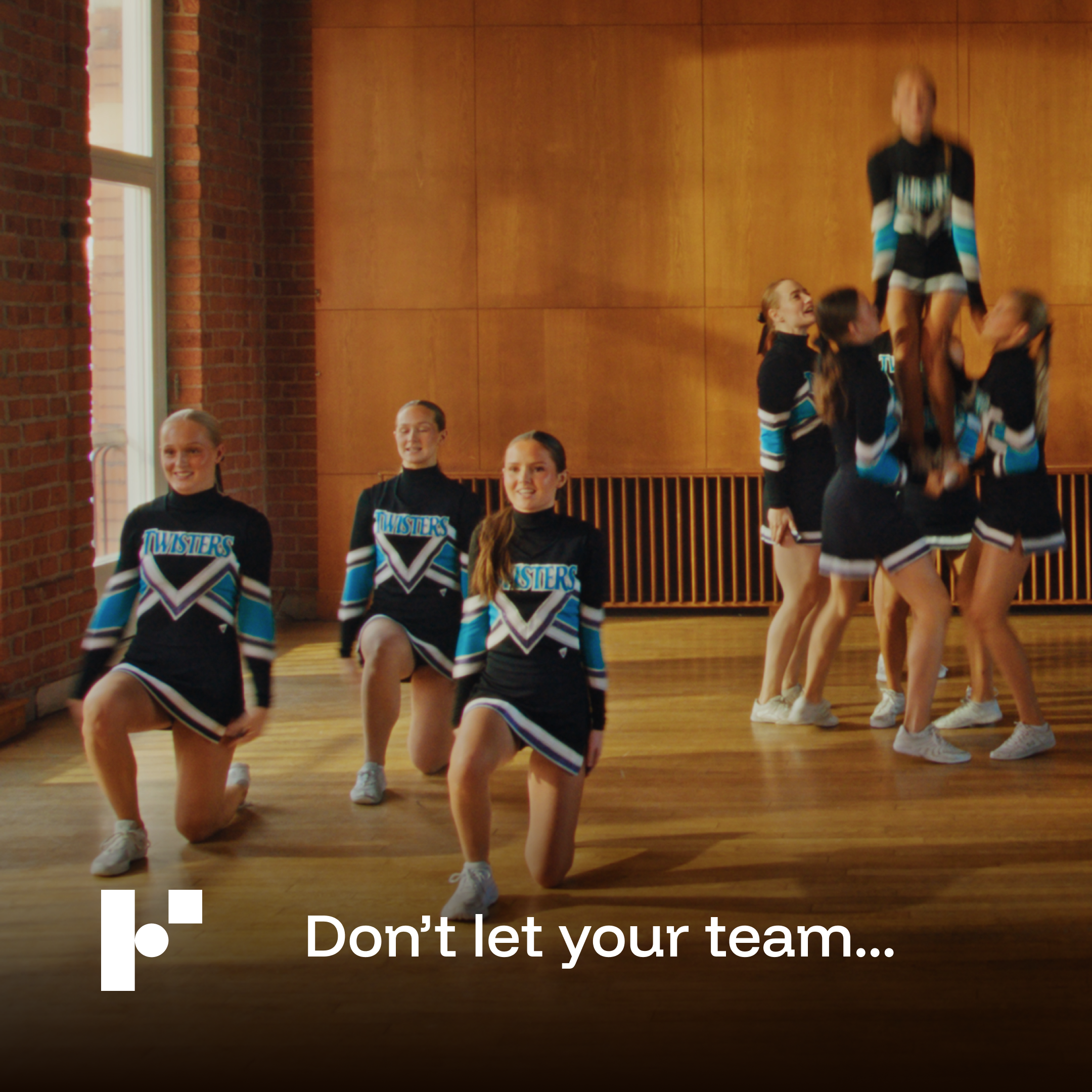 Girls in cheerleading uniforms performing stunts indoors with a brick wall and large window in the background.