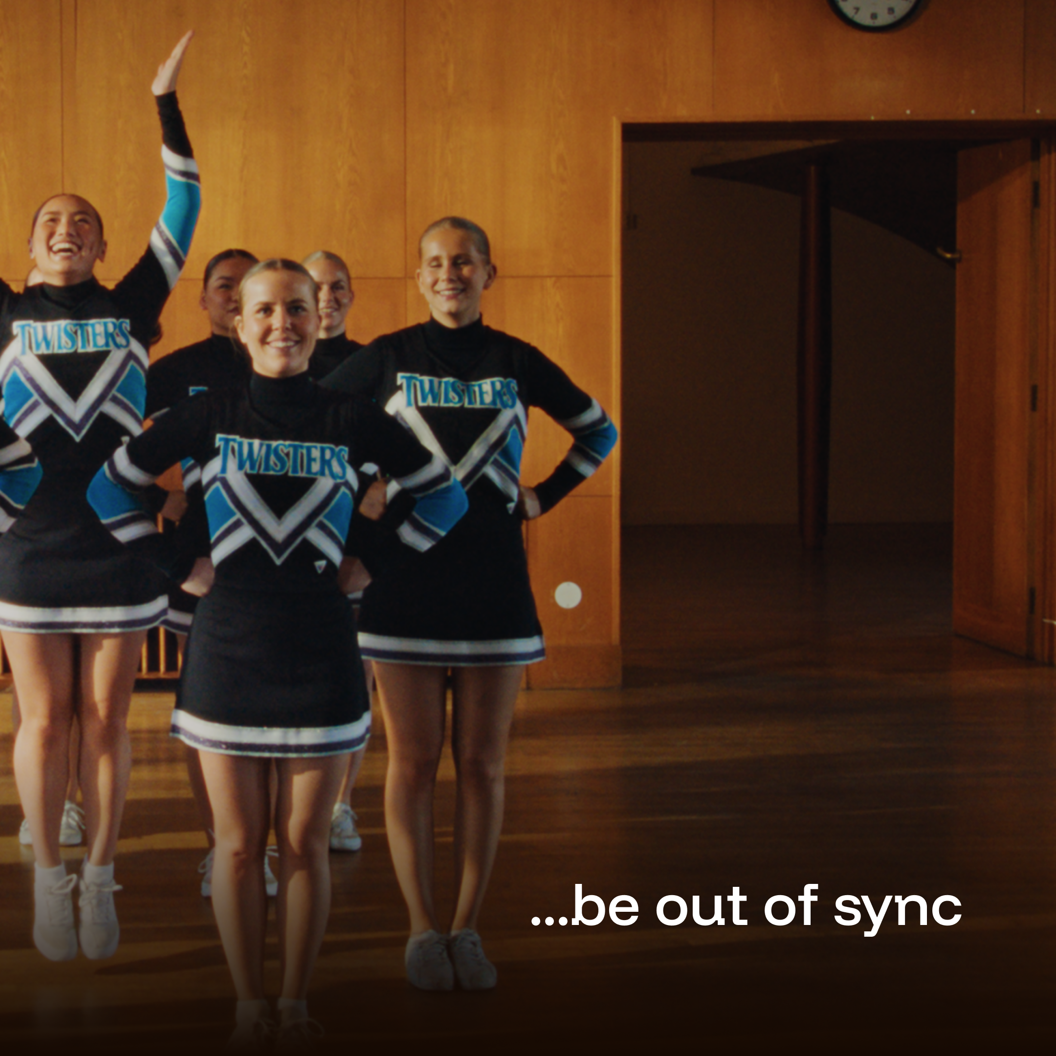 Cheerleaders in black, blue, and white uniforms with 'Twisters' written on them, standing indoors on a wooden floor, some smiling, one raising her arm, in a room with wood-paneled walls and a clock, with the caption '...be out of sync'.