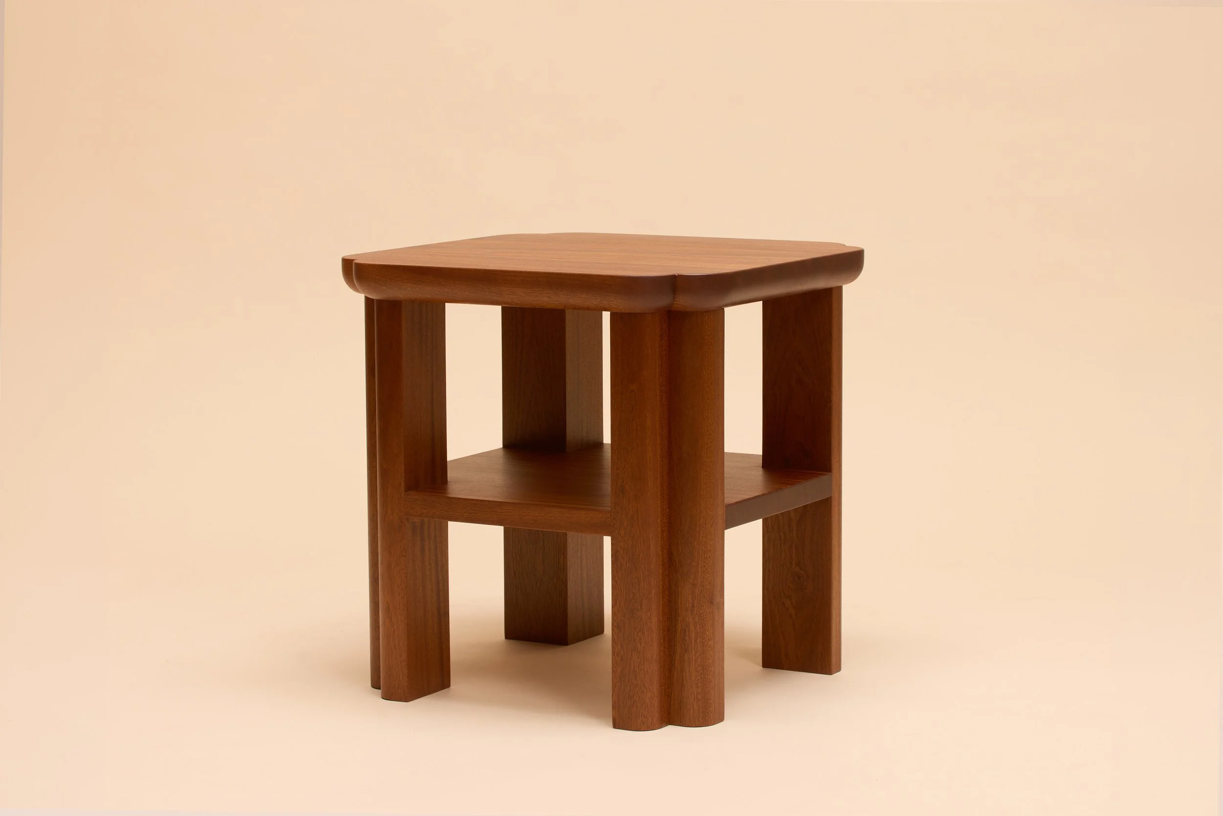 A side table in sapele with a integrated shelf, top has four rounded corners that meet the curvature of thick legs, against a beige background.