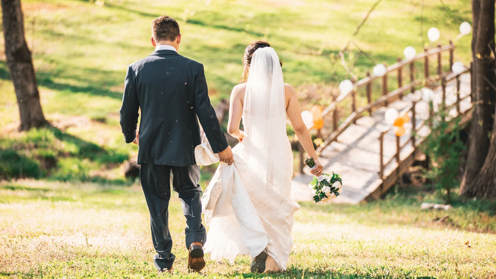 Bride and TGroom walk through sunny woods towards a wooden bridge