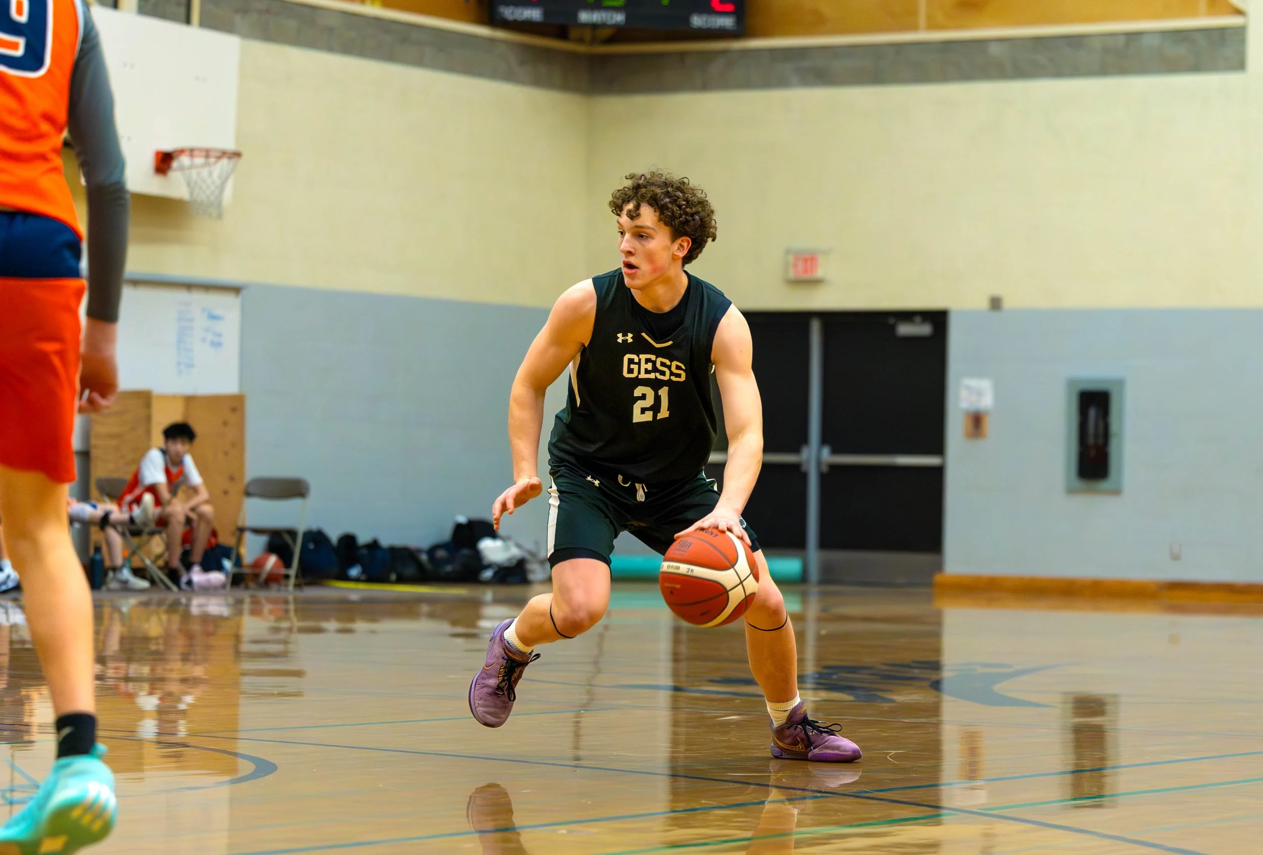 A young male basketball player with curly hair, wearing a black GESS jersey with the number 21, dribbling a basketball on an indoor court during a game.