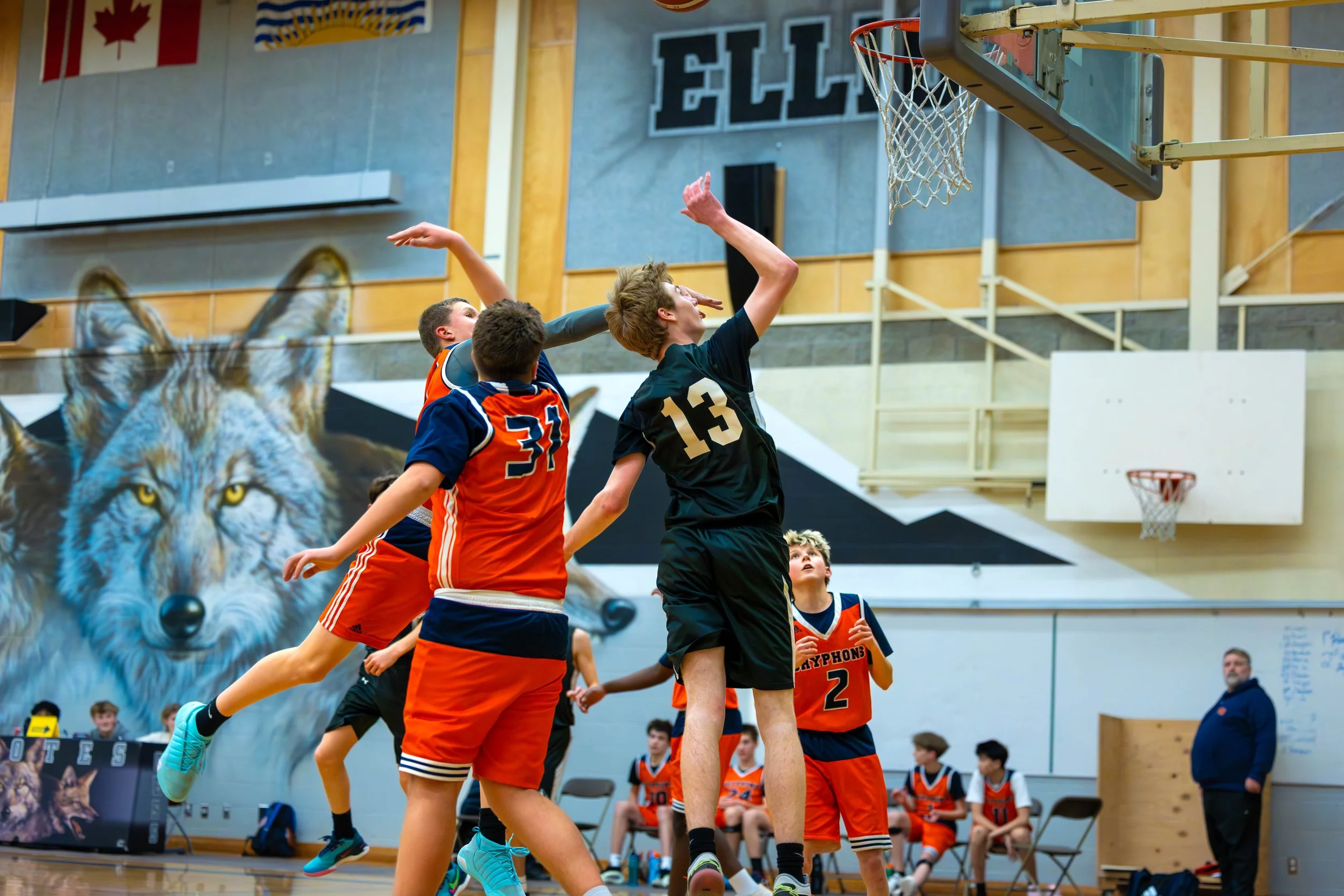 Young boys playing basketball indoors, with one player jumping to block a shot and others watching or preparing to rebound, against a backdrop of sports decor including a large wolf mural.