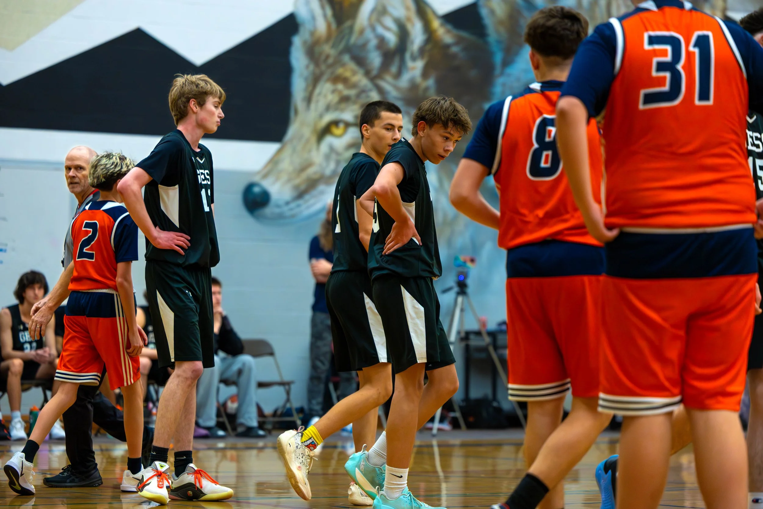 Teenagers in basketball uniforms standing on an indoor court, some appearing to listen to instructions, with a mural of a wolf on the wall in the background.