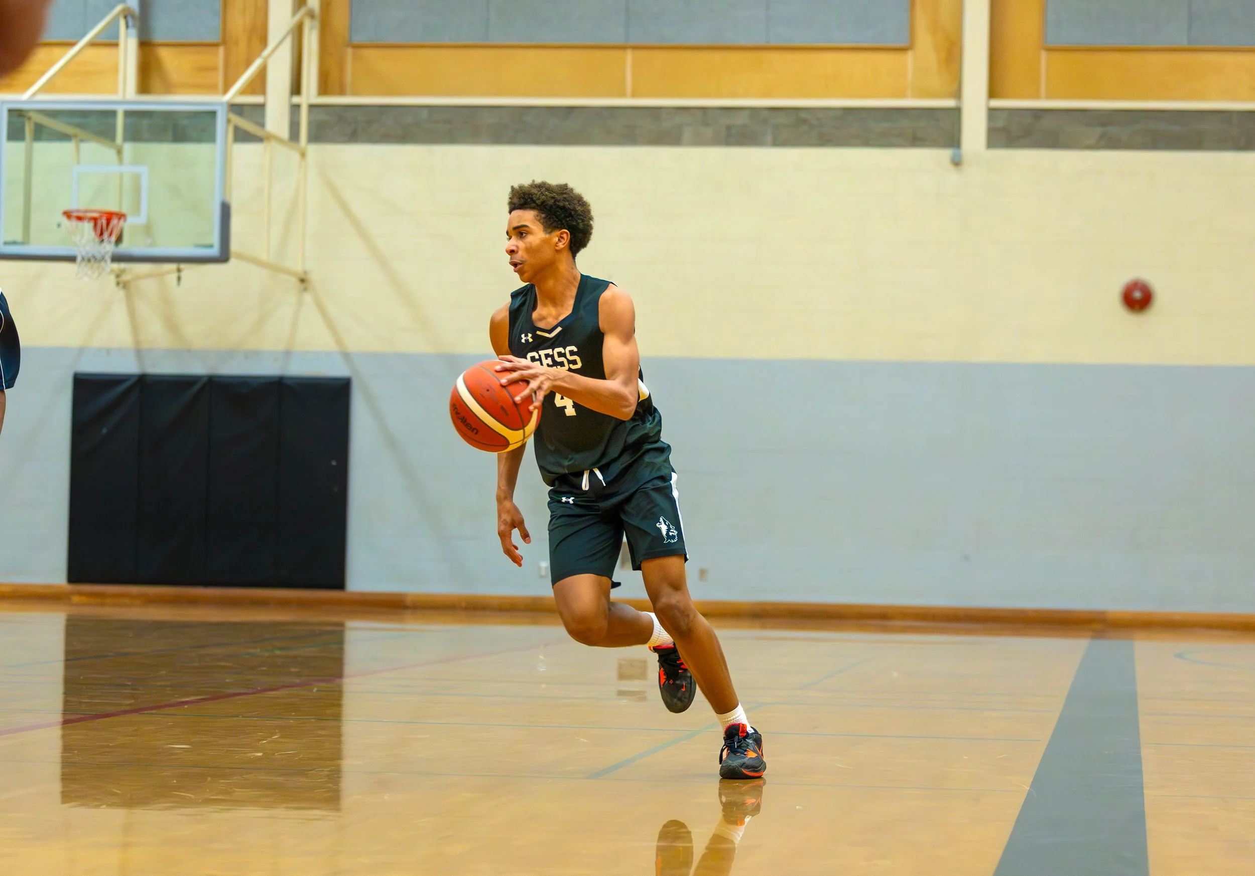A young male basketball player practicing on an indoor court, wearing a black jersey and shorts, dribbling a basketball.
