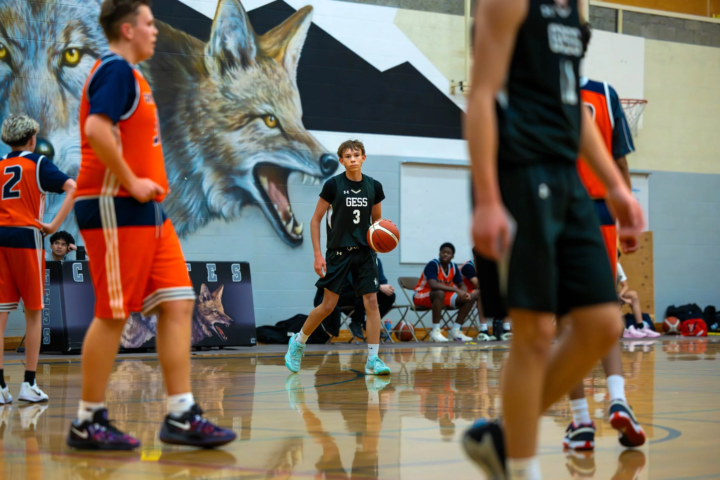 Young basketball player in a black uniform dribbling a basketball on an indoor court, with a mural of a wolf on the wall behind him, and other players and coaches present.