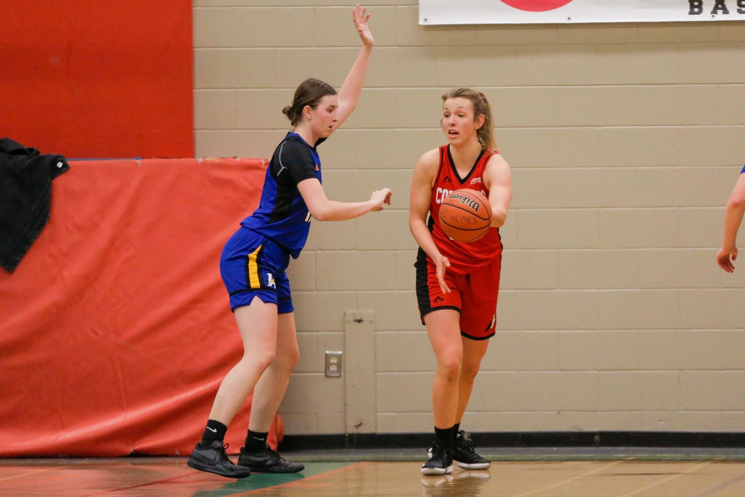 A girl in a red basketball uniform holding a basketball against a girl in a blue uniform trying to guard her during a game.