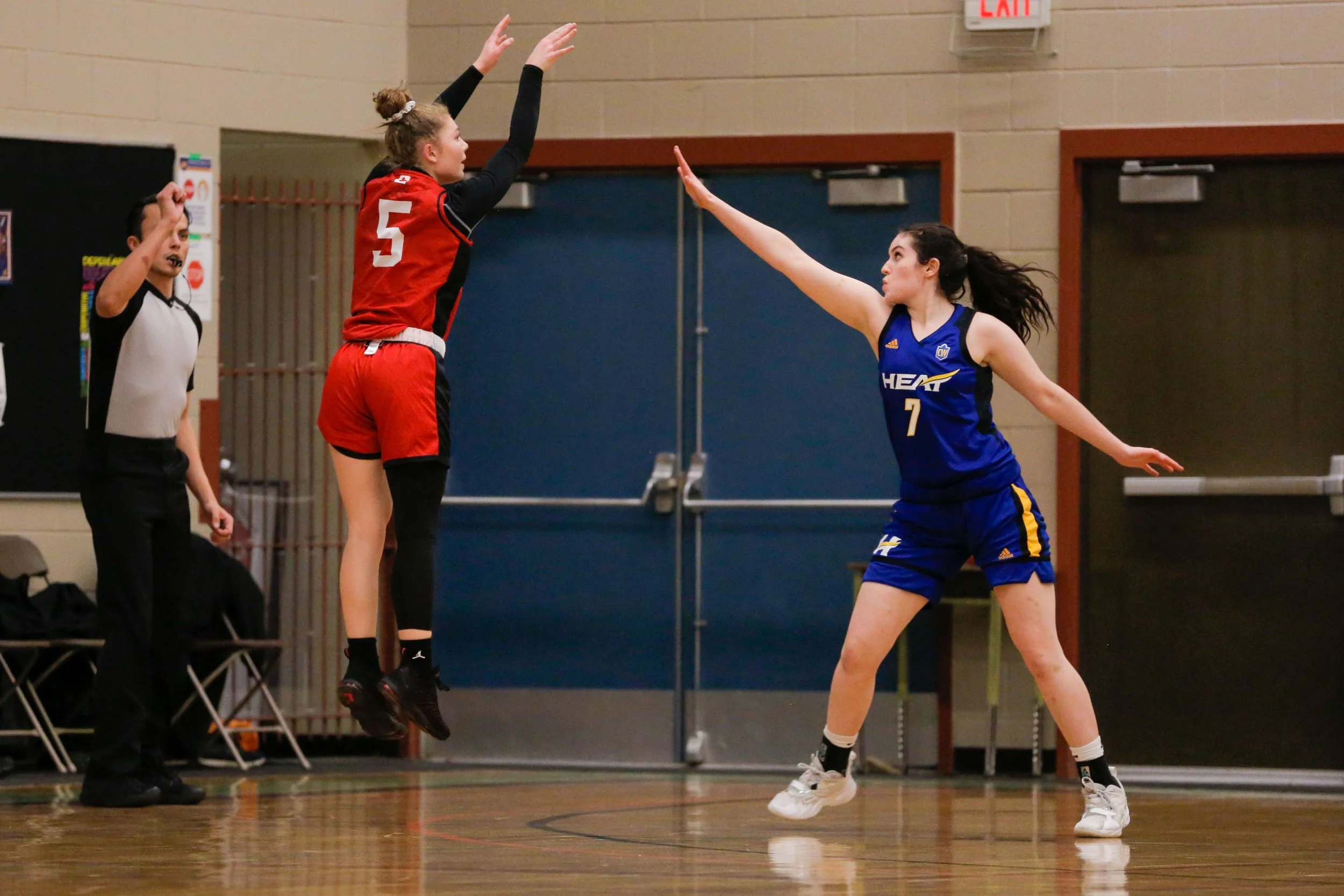 A basketball game in progress with a female player in a red and black uniform jumping to block a shot from a female player in a blue and yellow uniform. An official is standing nearby.
