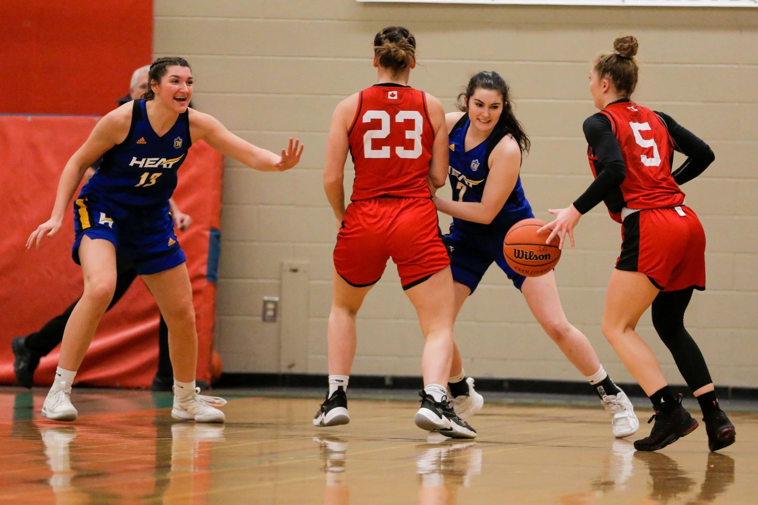Four female basketball players compete for the ball during a game. Two players wear red jerseys, and two wear blue jerseys. The player in blue with jersey number 14 is reaching for the ball, while the player in red with number 23 blocks her. The othe