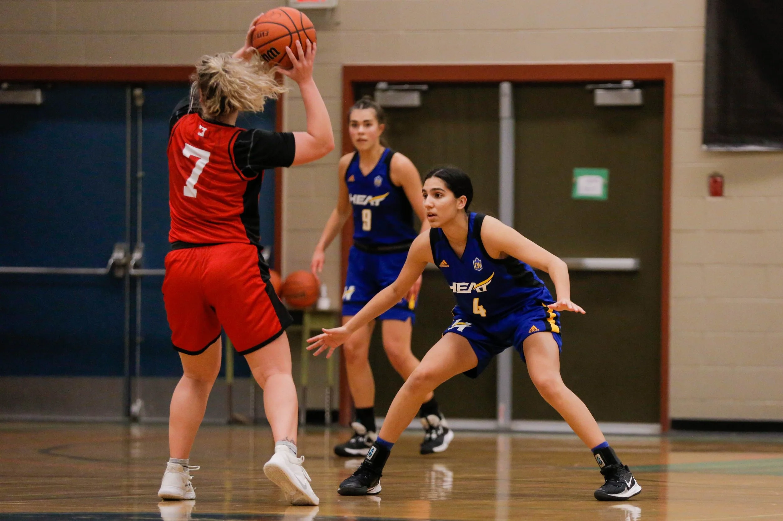 Two female basketball players in a game, one in a red and black uniform holding the ball above her head, while the other in a blue uniform defends with arms outstretched.