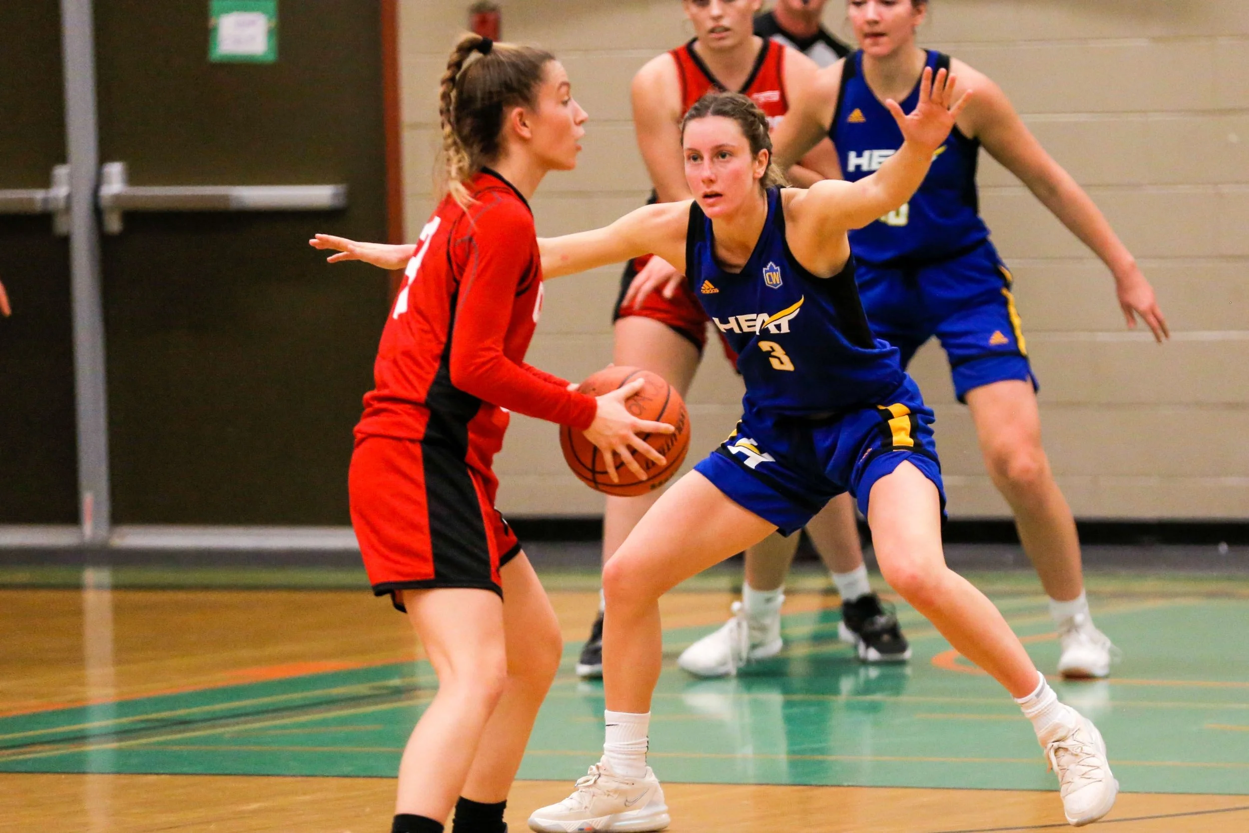 A women's basketball game with one player in red holding the ball while defending against a player in blue with arms outstretched. Two more players in red and blue are in the background.