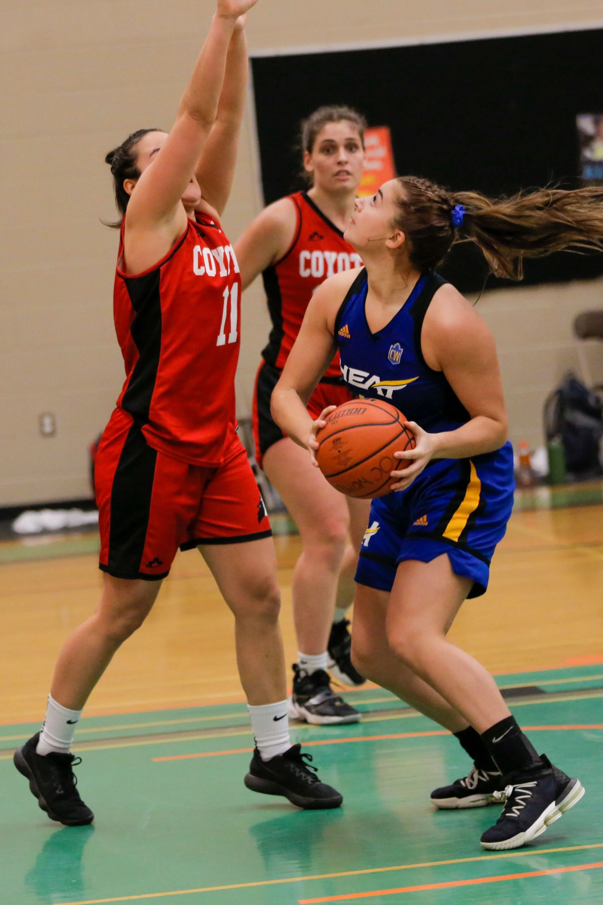 A women’s basketball game showing three players: two in red jerseys attempting to block a woman in a blue jersey who is holding the basketball, on a basketball court indoors.