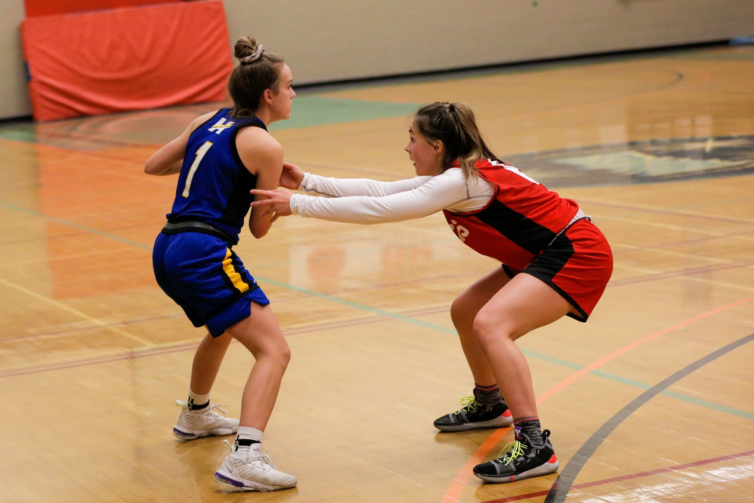 Two female basketball players are fighting for possession of the ball on an indoor basketball court. The player in blue is trying to hold onto the ball while the player in red is reaching for it.