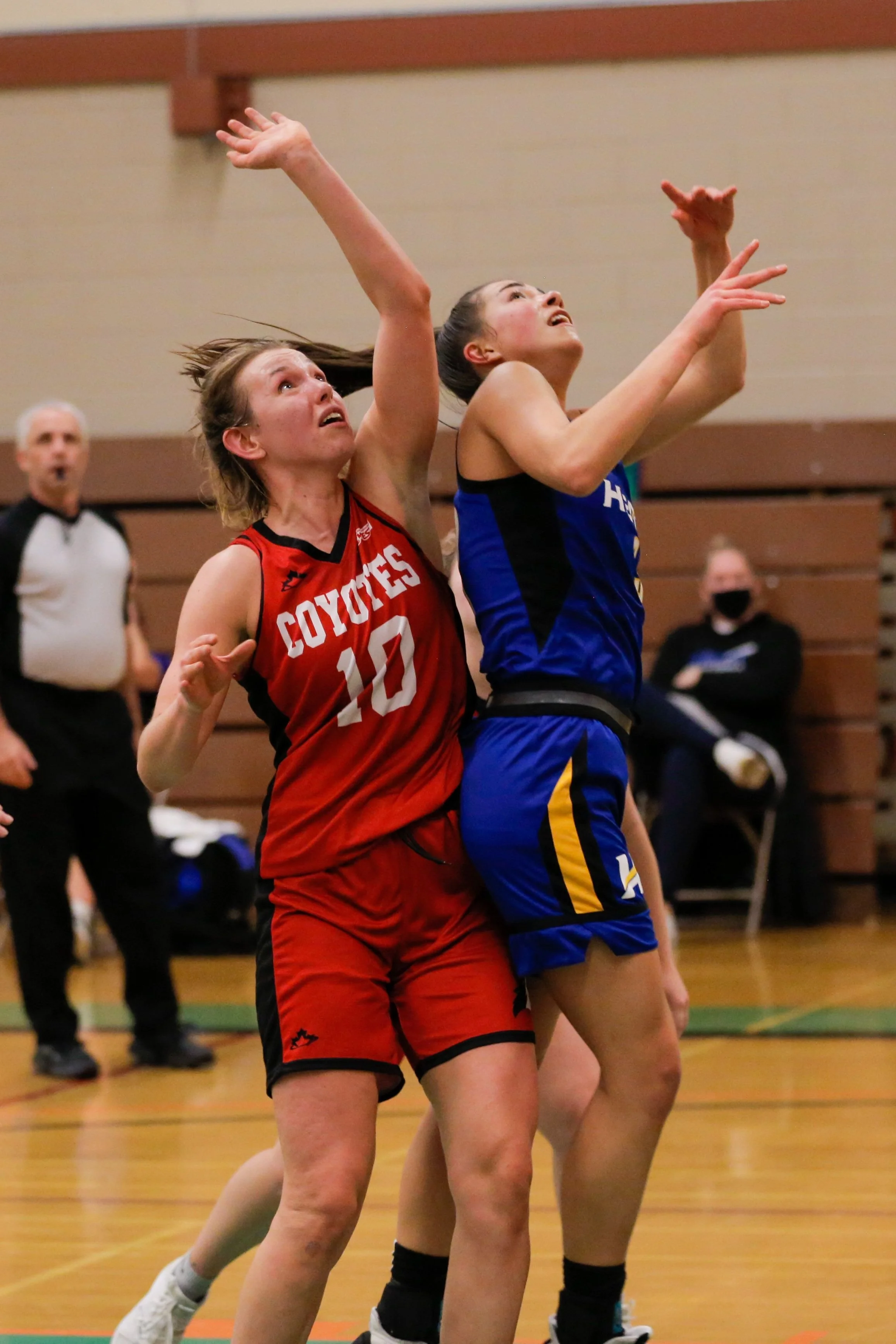 Two female basketball players jumping for the ball during a game, one in a red jersey with 'Coyotes' and the number 10, and the other in a blue jersey, in an indoor gymnasium.