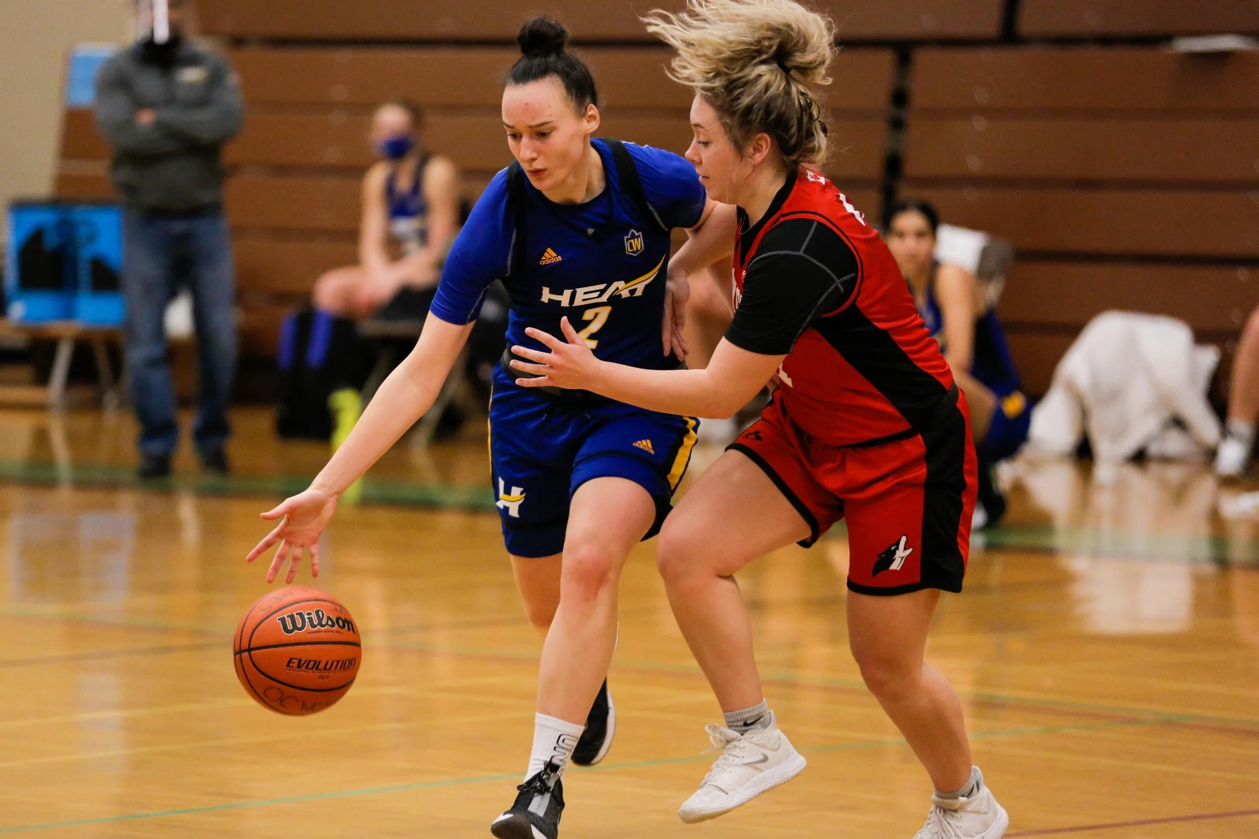 Two women playing basketball on an indoor court, one in a blue uniform and the other in red, battling for possession of the ball.