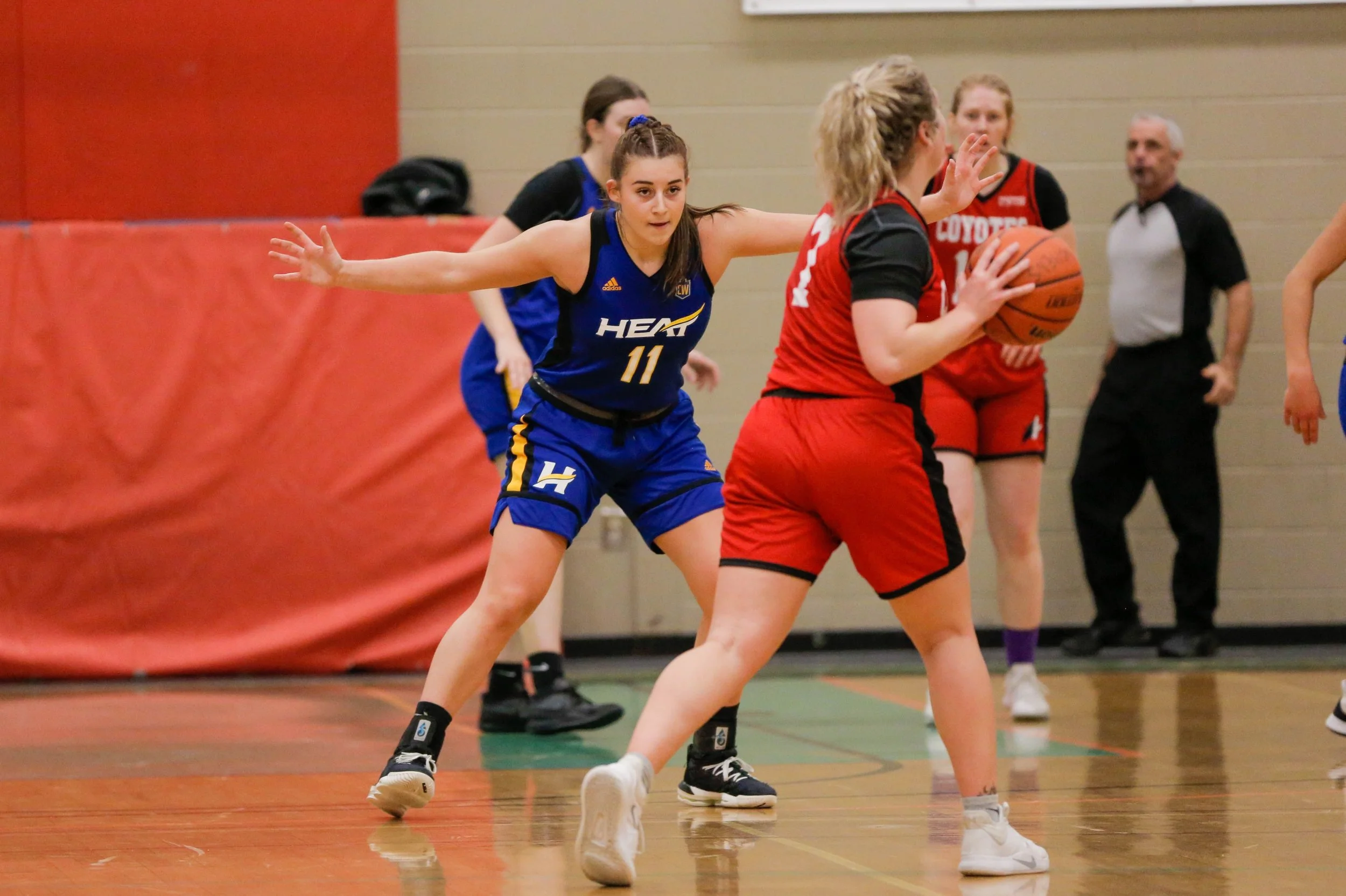 A women's basketball game with players in blue and red uniforms, one player in red holding a basketball while being guarded by a player in blue with arms outstretched.