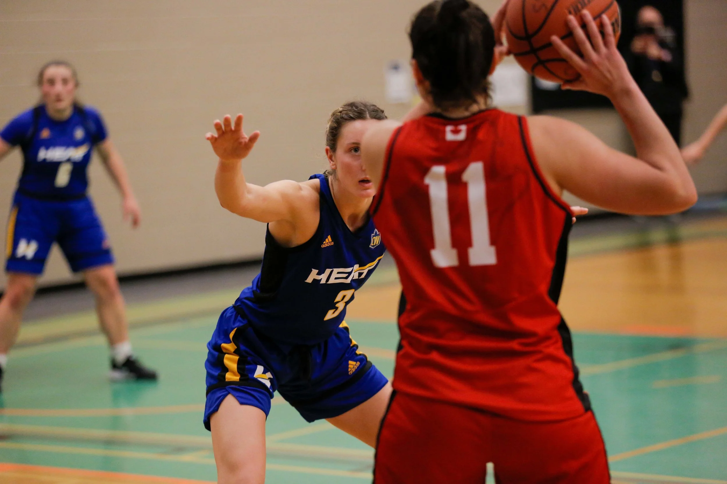 A female basketball player in a blue uniform guarding an opposing player in a red uniform who is holding a basketball in a gymnasium.