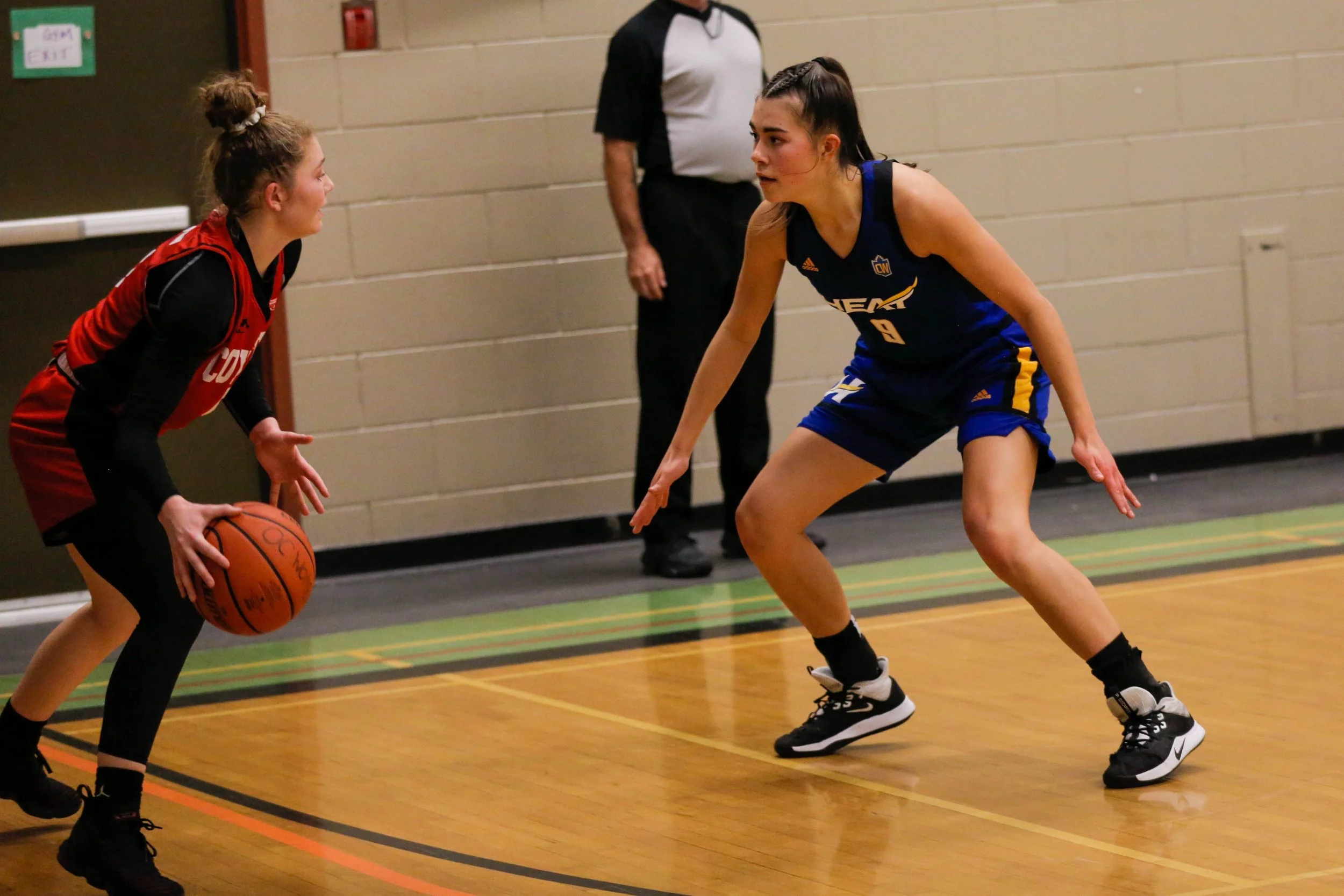 Two female basketball players are in a defensive stance on the court, with one player dribbling the ball and the other player defending her. An official is standing in the background.