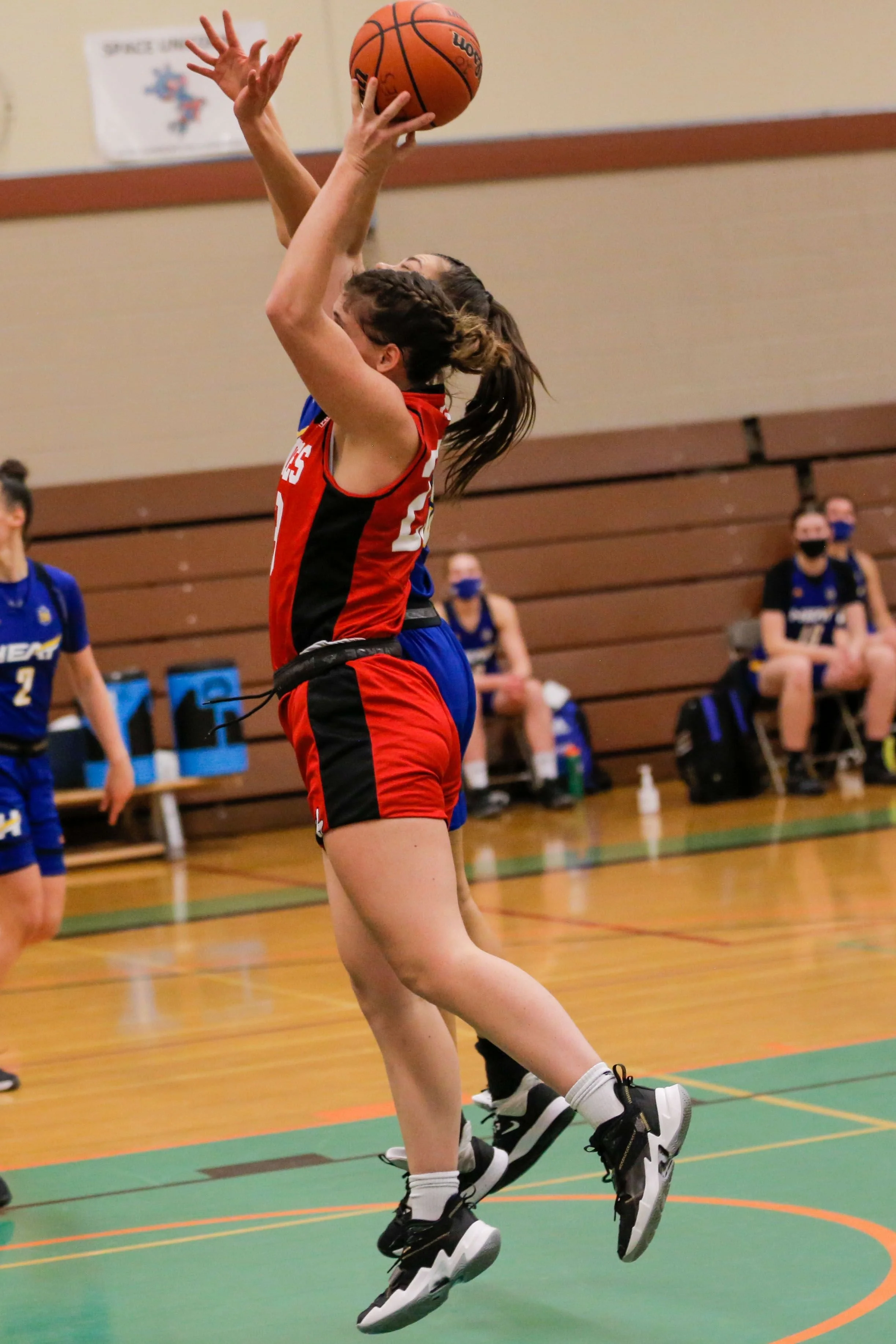 A girl in a red and black basketball uniform jumps to make a shot during a game, with other players and seated spectators in the background.
