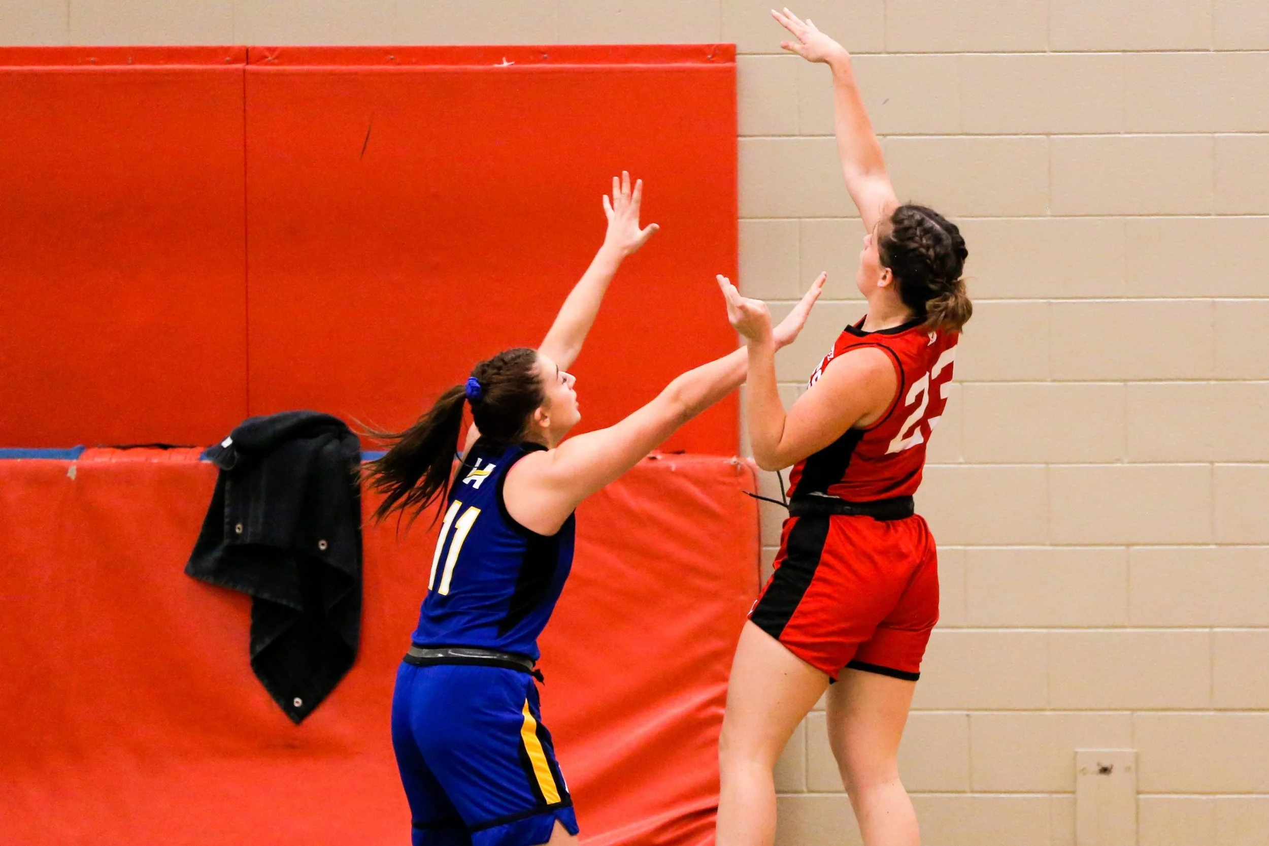 Two female basketball players, one in a blue uniform and the other in a red uniform, are jumping with arms extended toward each other, contesting a play near a gymnasium wall.