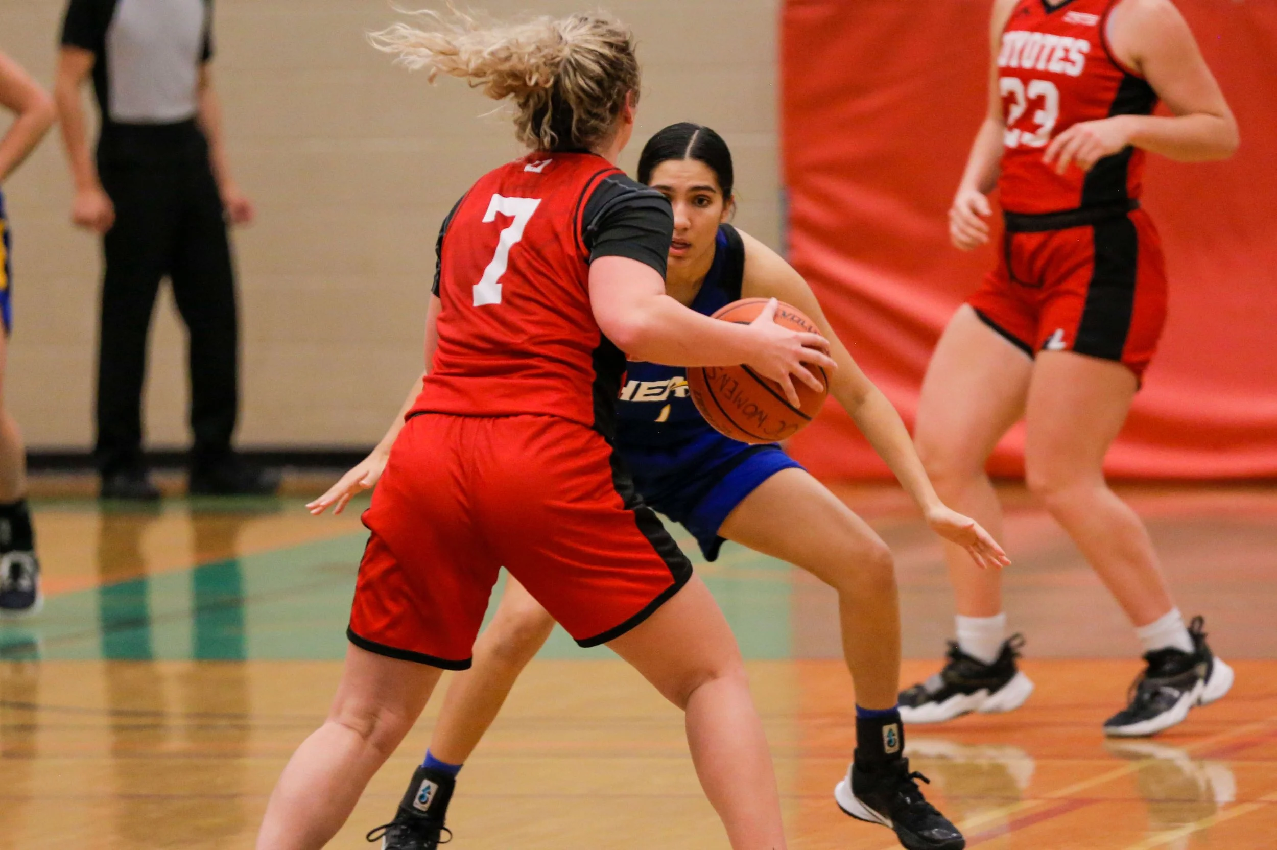 Two female basketball players compete for the ball during a game, one in a red uniform and the other in a blue uniform.