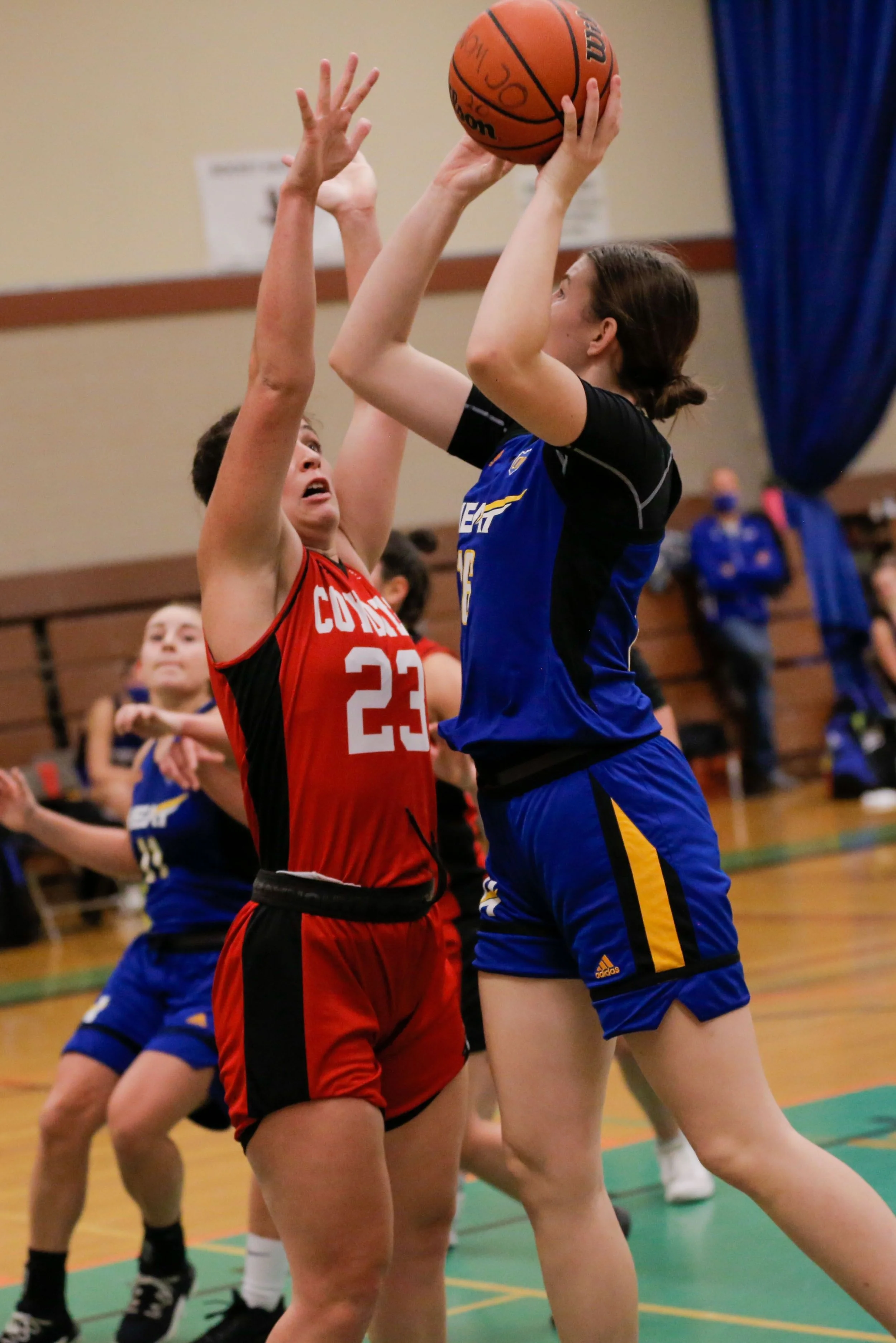 Two female basketball players are competing for the ball near the basket, one in a red jersey with number 23 and the other in a blue jersey. The player in blue is jumping to shoot or pass the ball, while the player in red is attempting to block or de
