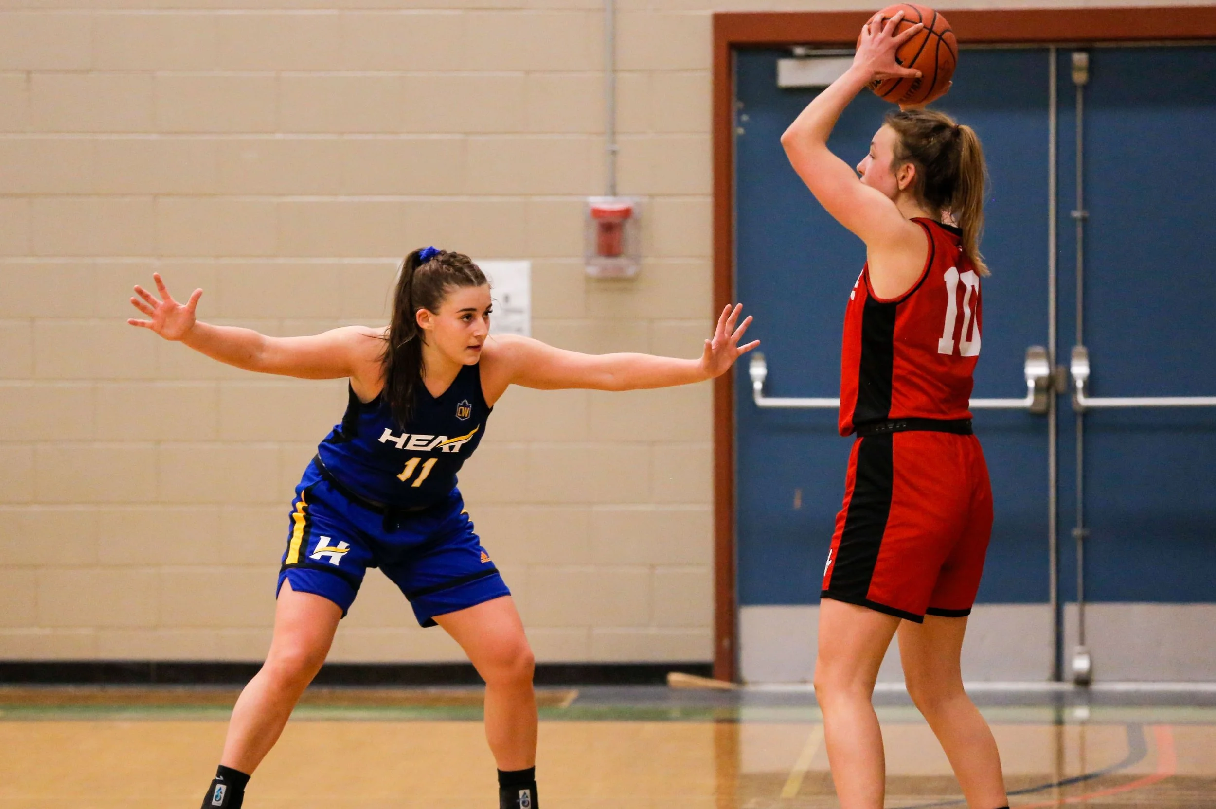 Two female basketball players in a gymnasium, one in a blue uniform with the number 11, defending against another player in a red uniform, who is holding a basketball above her head.