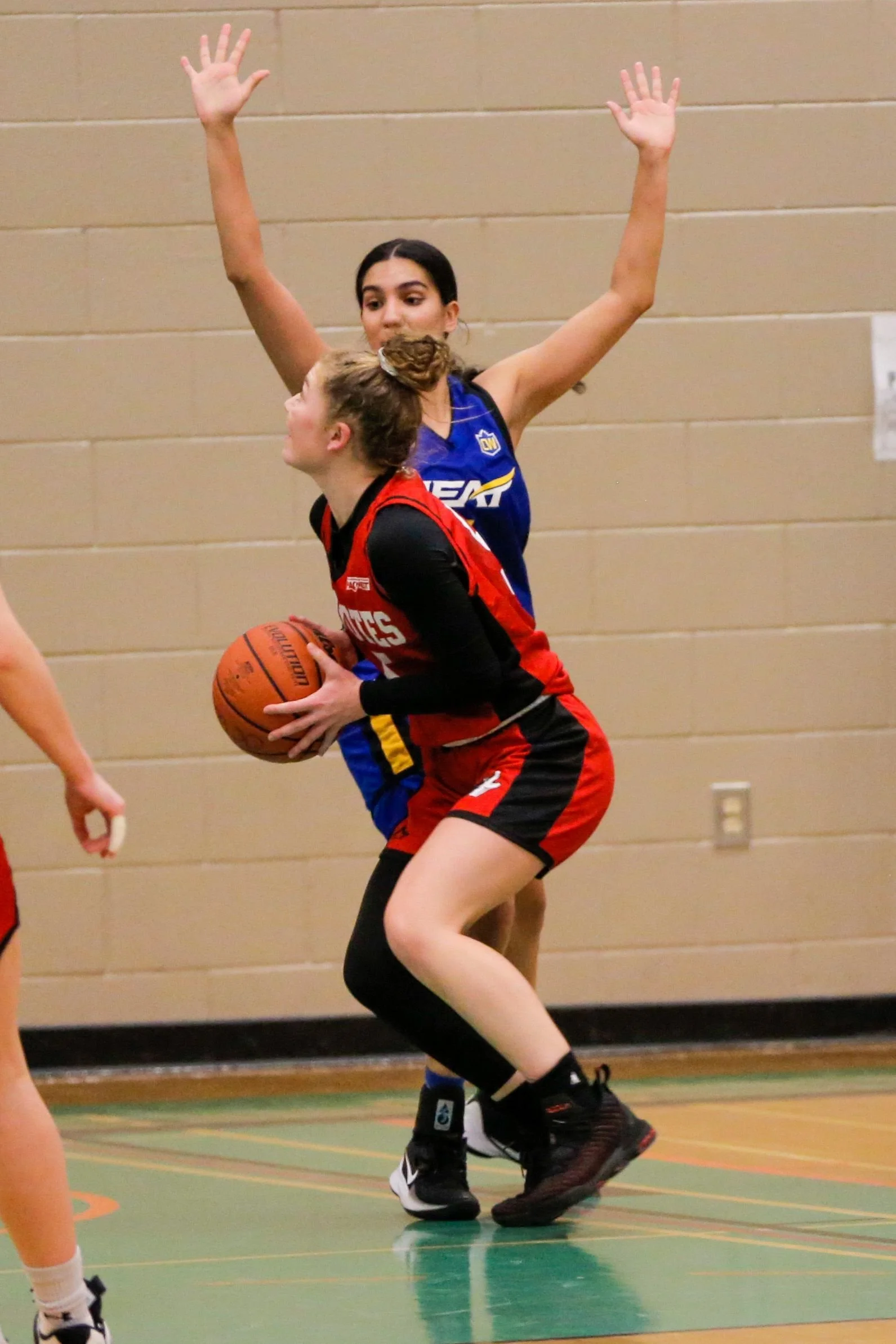 Female basketball player in red uniform holding a basketball, trying to pass or shoot, with an opponent in blue defense behind her, inside a gymnasium.