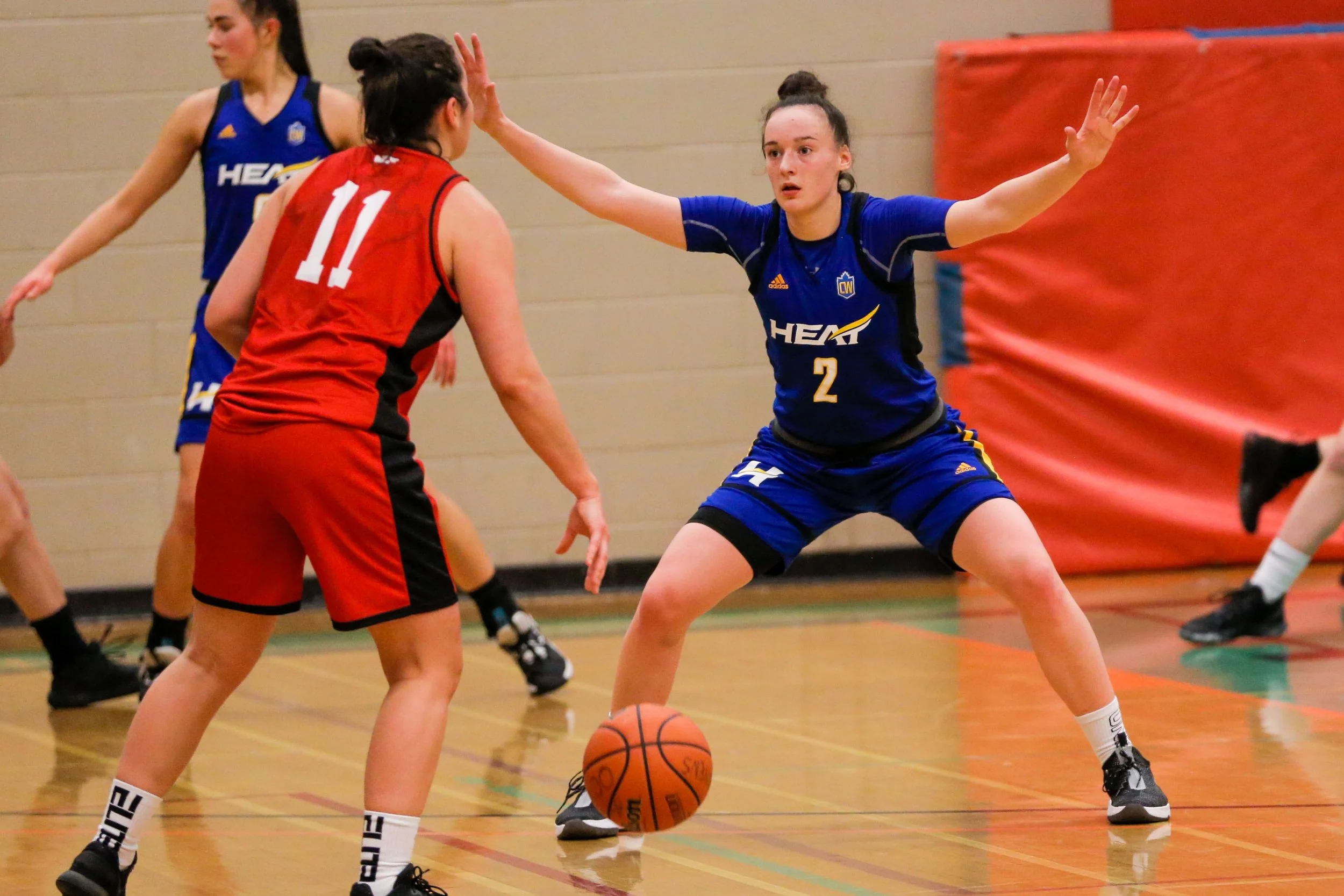 Two female basketball players compete for possession of the ball during a game, with one wearing a red jersey numbered 11 and the other in a blue jersey numbered 2, on an indoor court.