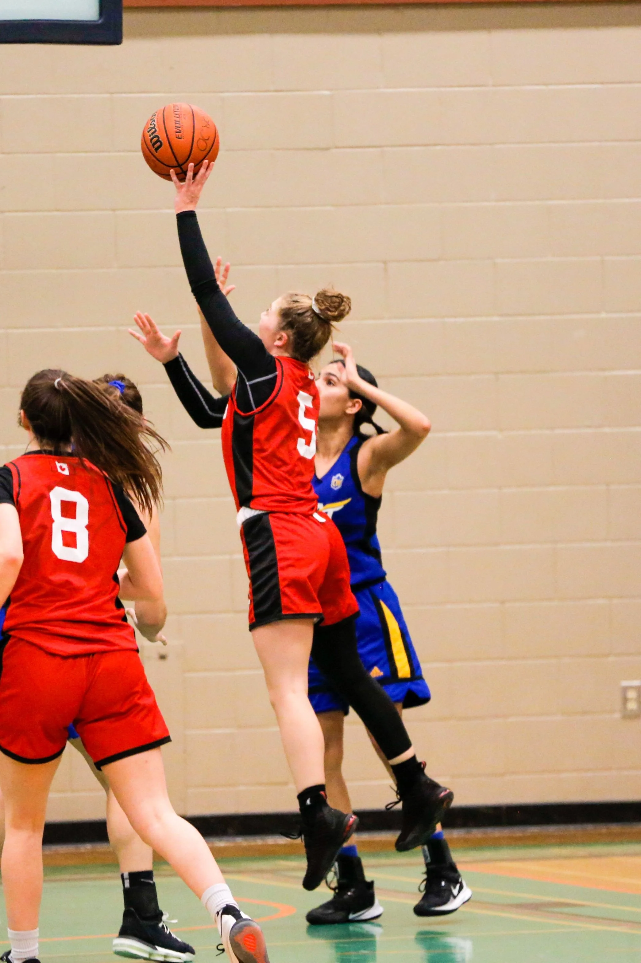 Girls playing basketball, with one girl jumping to shoot or block the ball, wearing a red and black sports uniform, on an indoor court.