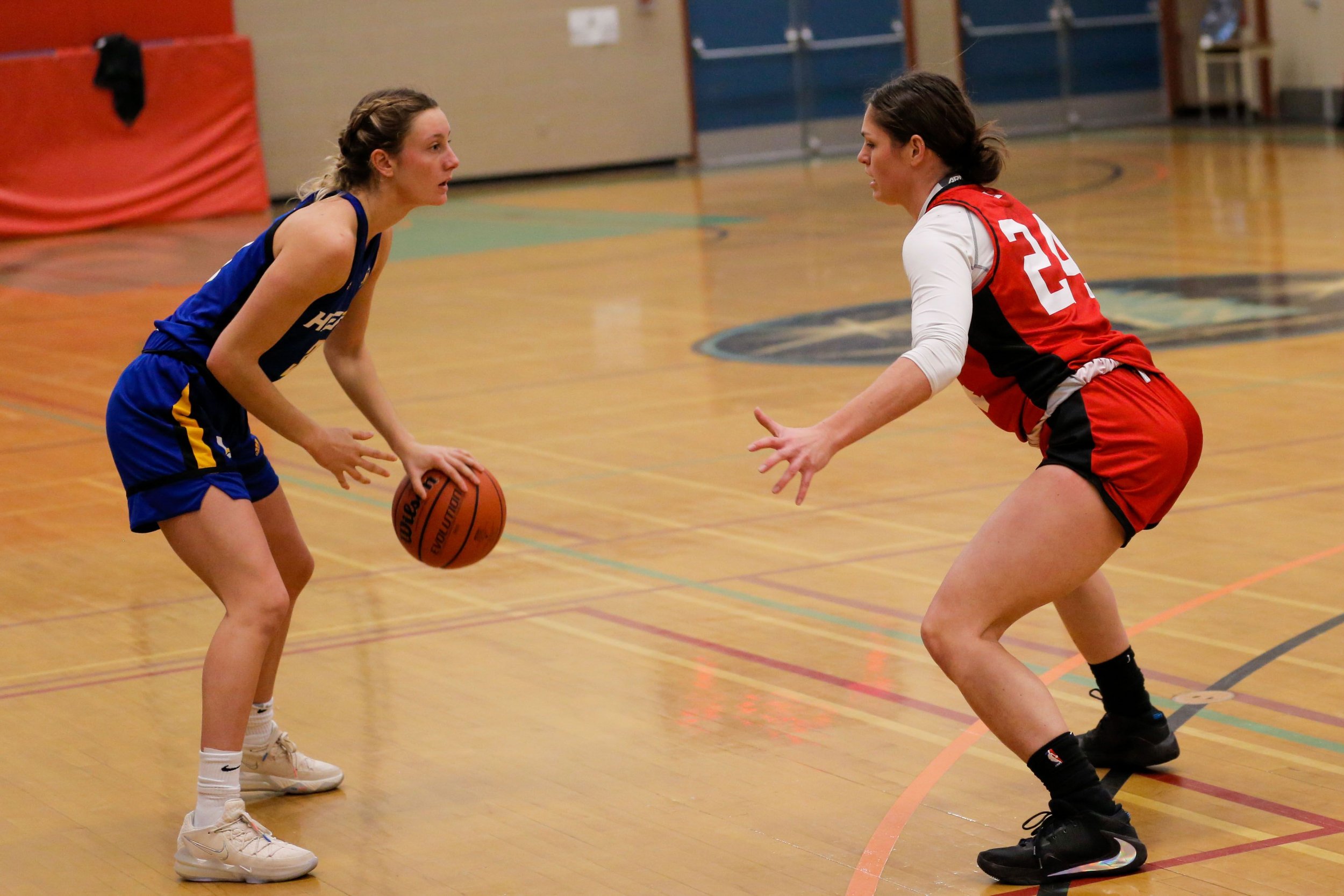 Two female basketball players on an indoor court, one in a blue uniform holding a basketball, and the other in a red and white uniform defending.