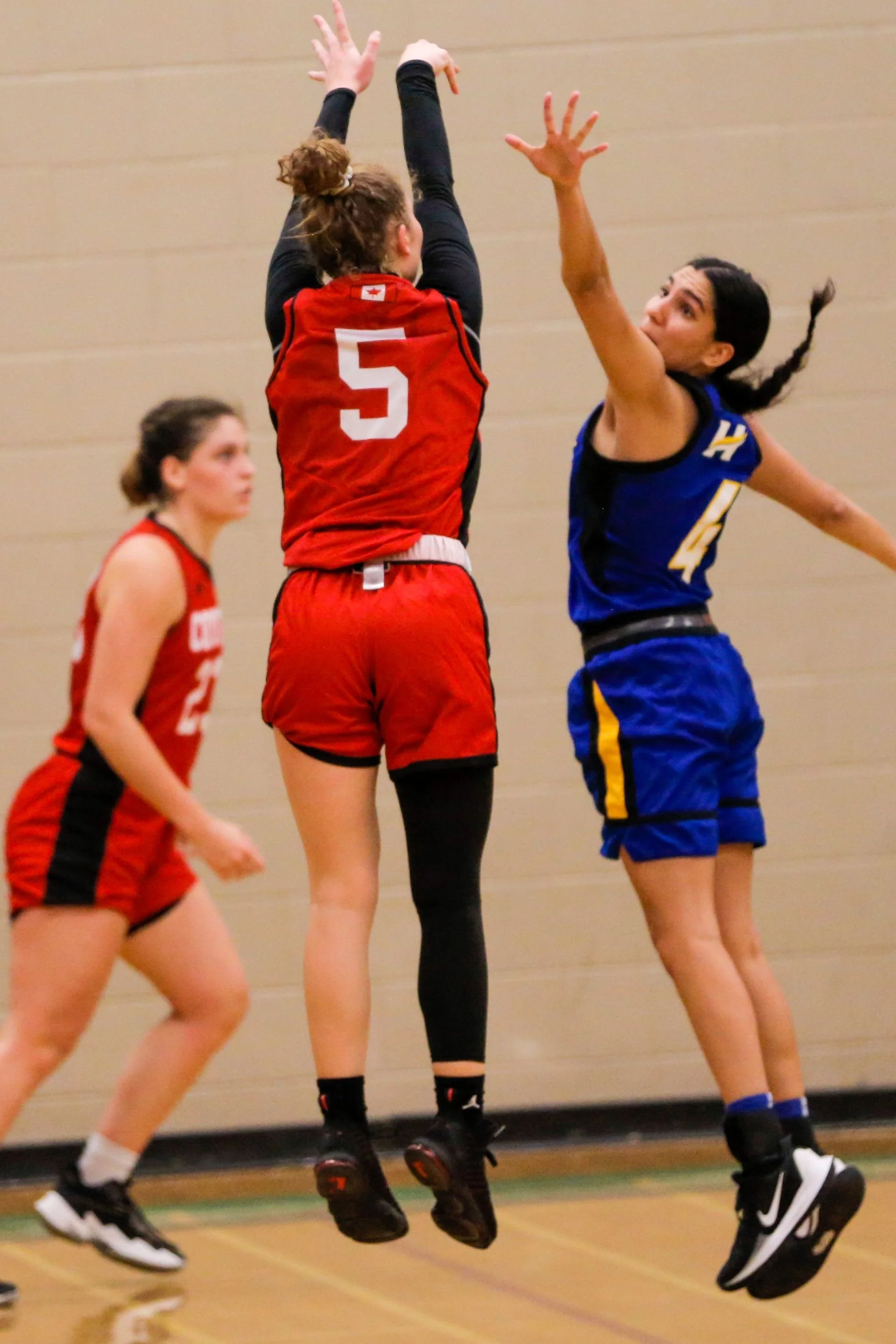 Two female volleyball players, one in red and one in blue, jump to block or spike the ball during a game, with a third player in red in the background.