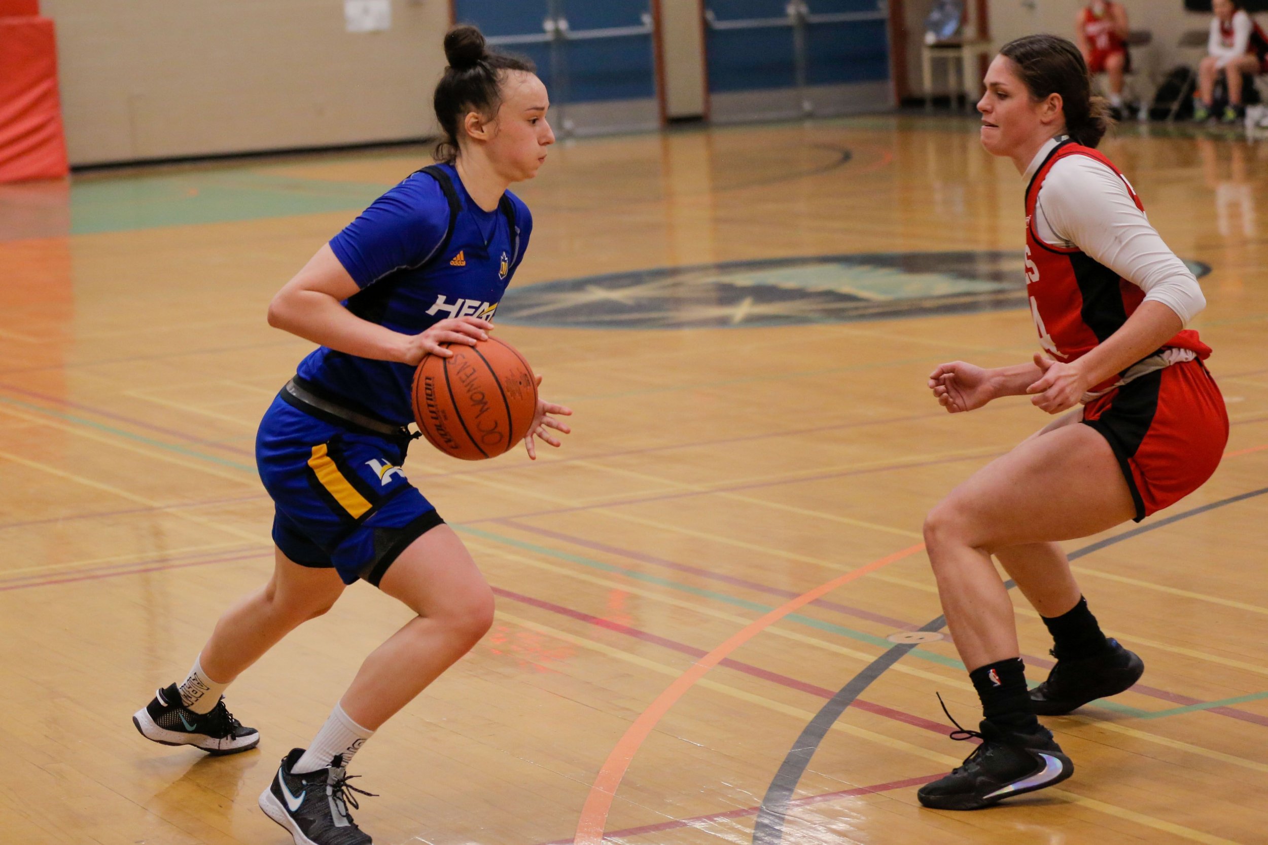 Two female basketball players are on a wooden court; one in a blue uniform with the ball, the other in a red and black uniform defending.