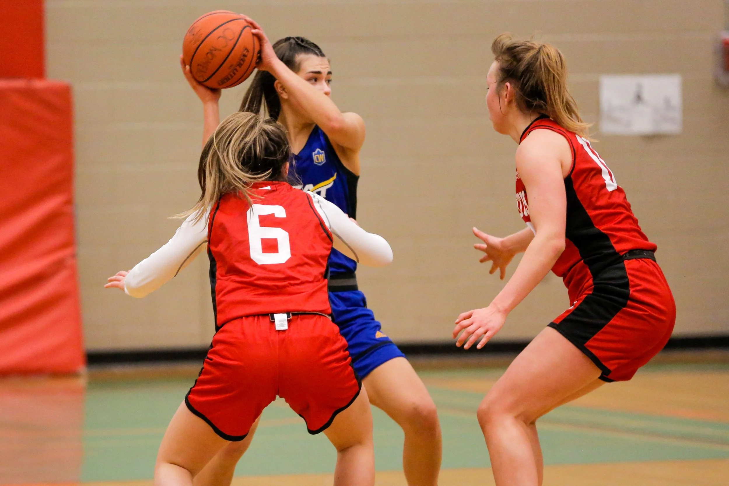 Three female basketball players in action during a game, with one in a blue jersey holding a basketball and two in red jerseys defending.