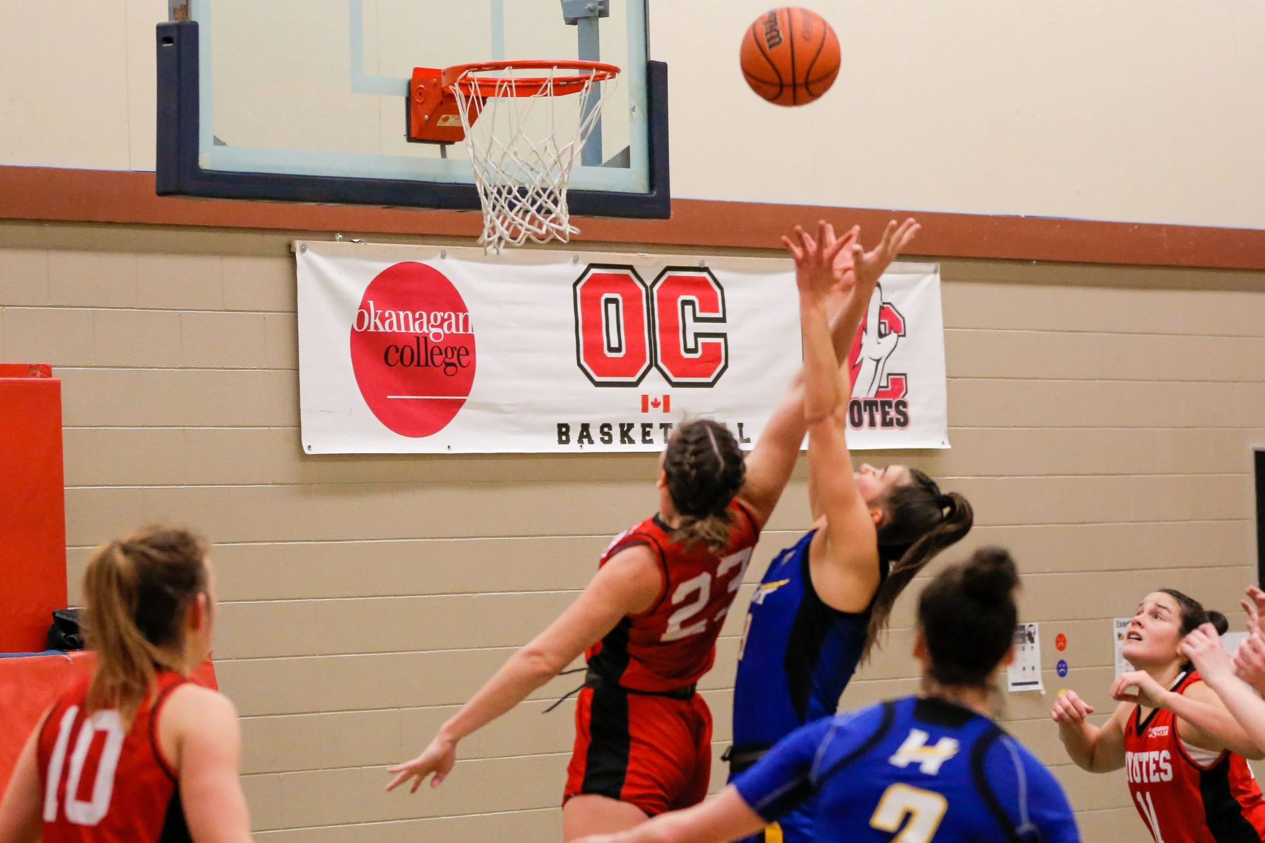 Two female basketball players, one in red jersey number 23 and the other in blue jersey, jump to block a shot near the basketball hoop during a game, with a banner for Okanagan College in the background.