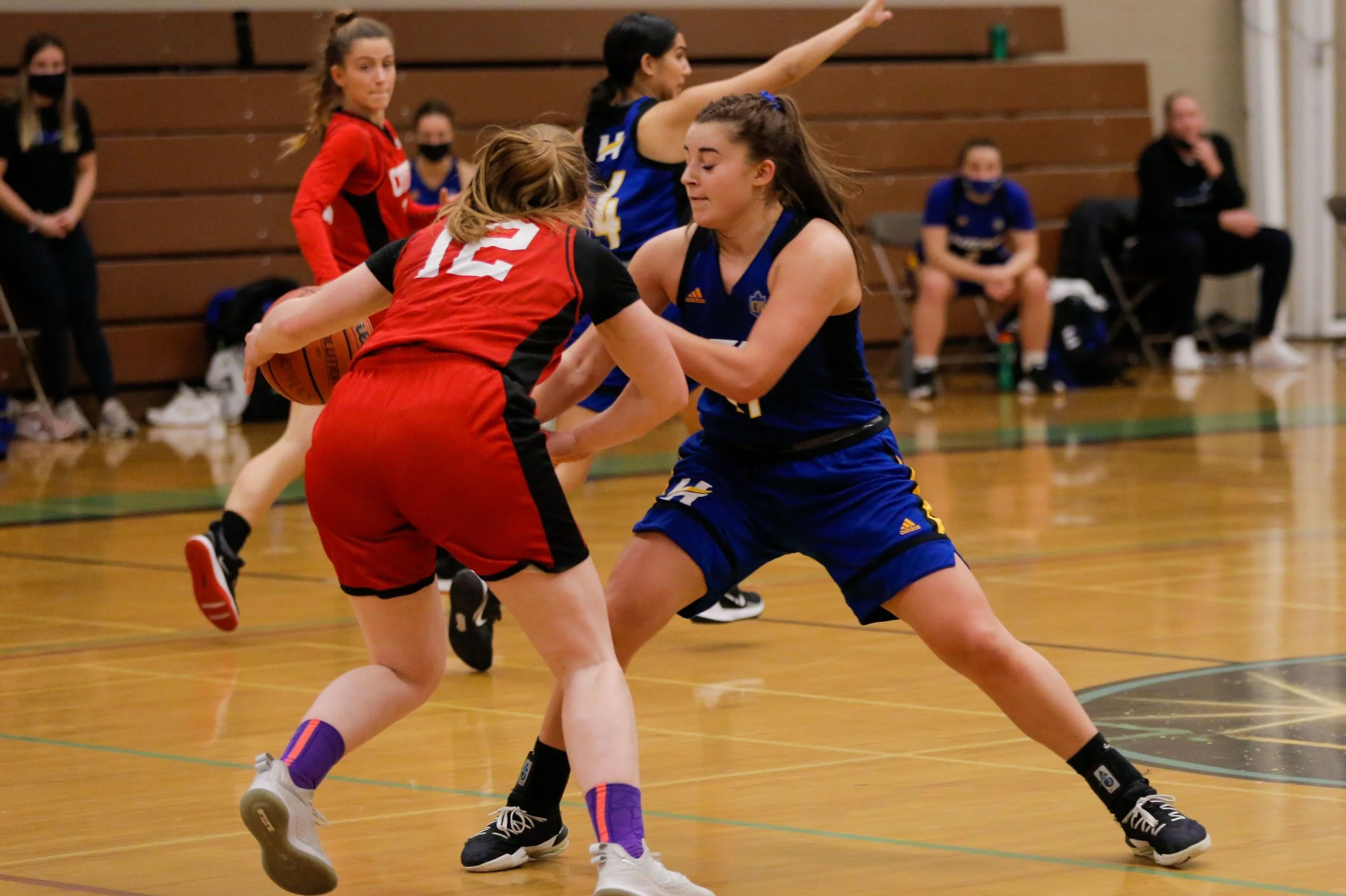 Two female basketball players, one in a red uniform and the other in a blue uniform, are playing defense and trying to steal the ball during a game. The player in red is holding the basketball, and the player in blue is reaching to block or steal it.