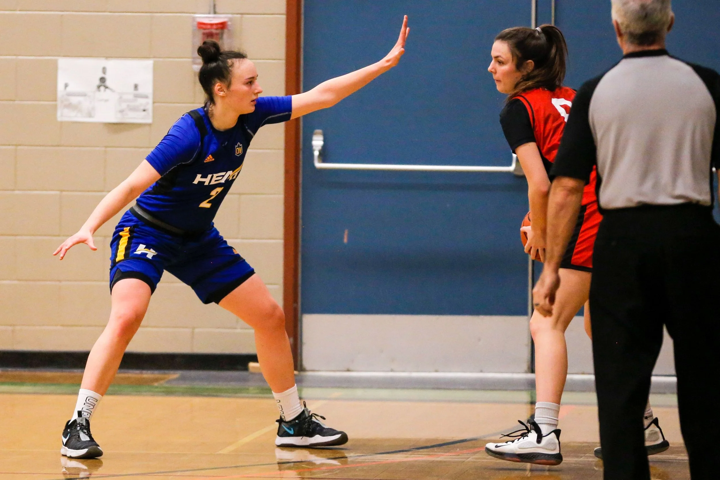Two women playing basketball during a game, one in a blue uniform and the other in a red and black uniform, with a referee observing nearby.